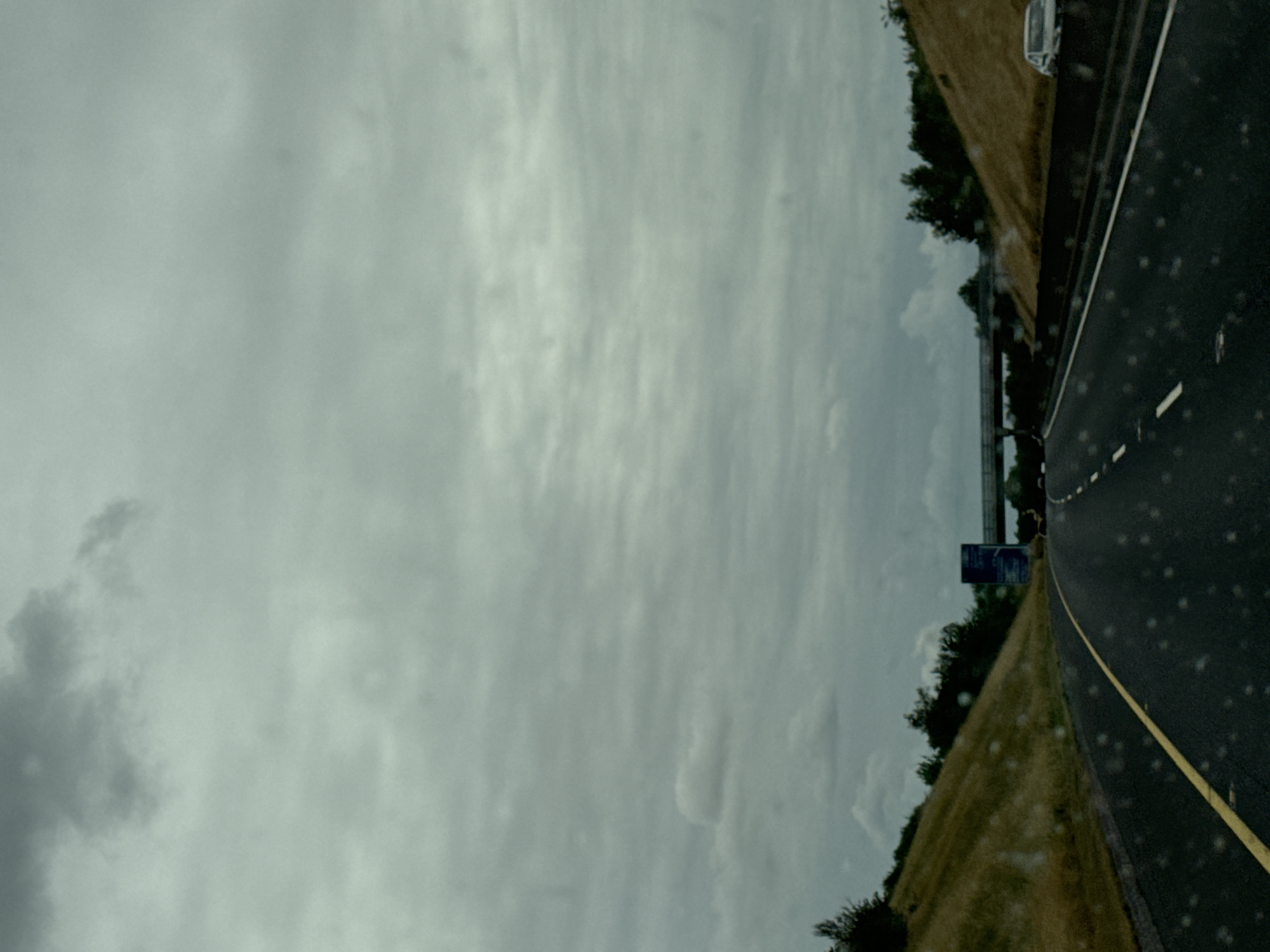 The image depicts a highway scene on a cloudy day. The perspective is from inside a vehicle, looking forward onto a wet asphalt road with a yellow line dividing the lanes. On either side of the road are grassy embankments. In the distance, an overpass spans the highway, and a blue signpost is visible on the left side of the road. A white car is seen on the right side of the highway. The sky is overcast with a heavy, grey cloud cover. There is no specific indication of the city of Nine Tree Hill, Ireland, or any people present in the image.