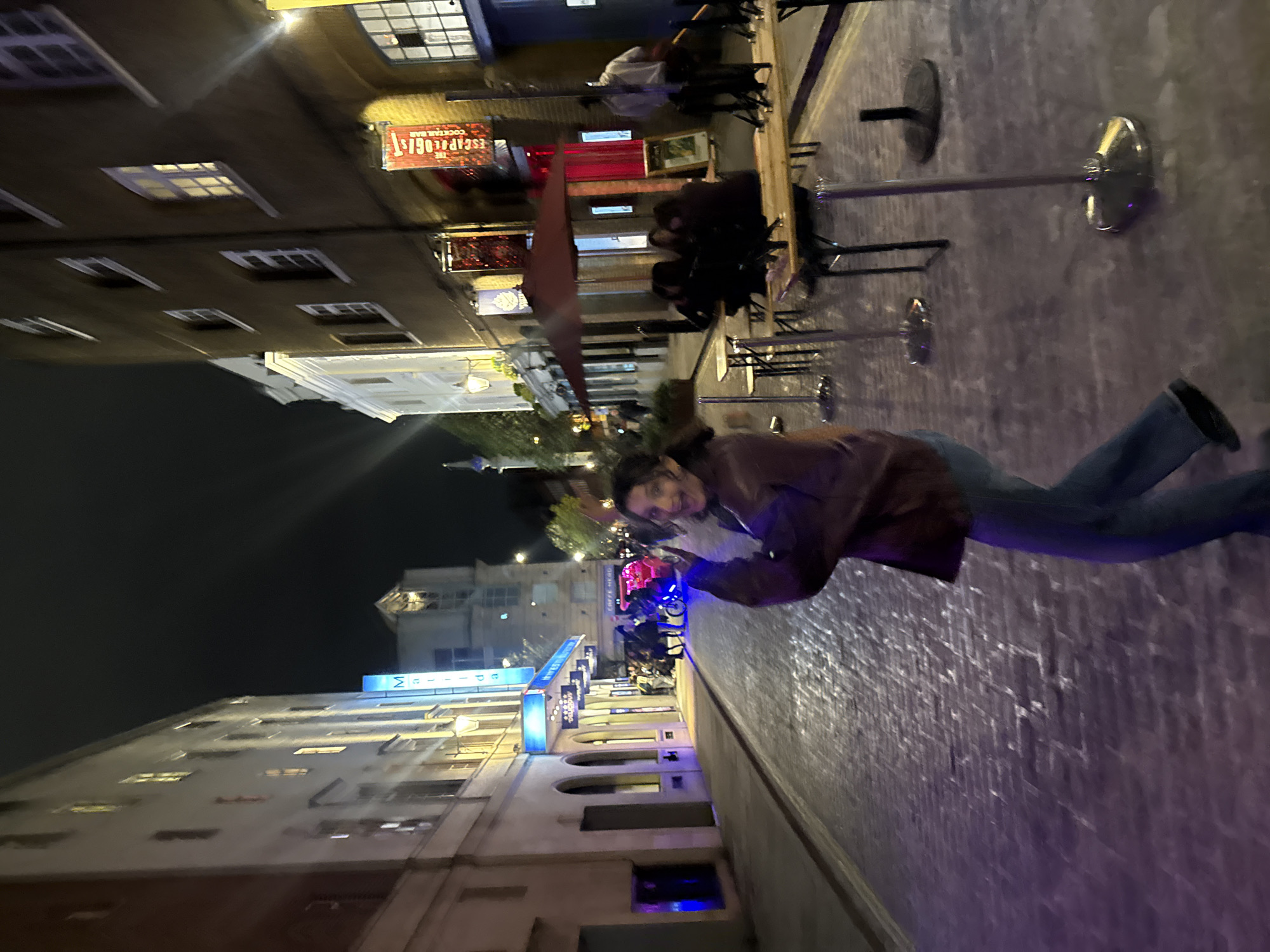 At night, a woman in a brown jacket and jeans stands on a cobblestone street in London, United Kingdom. She looks towards the camera, smiling. Behind her, people are seated at outdoor tables. The street is lined with buildings, some with neon signs. One sign reads "The Escape Lodge Cocktail Bar." Another sign is illuminated with blue neon light. The night sky is visible above the buildings.