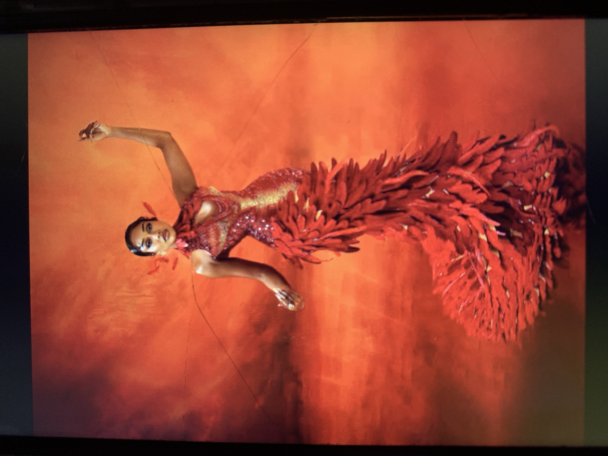 A woman in a red and gold shimmering gown with a large, feathered train is posed against a background that is predominantly red and orange. The woman is facing the viewer with her head tilted slightly to the left. Her right arm is raised above her head, and her left arm is extended to the side. The dress appears to be heavily embellished, particularly the train, which fans out behind her. The lighting emphasizes the texture and detail of the dress and the background. There is no information about the location being in New Stadium, Nigeria in this image.