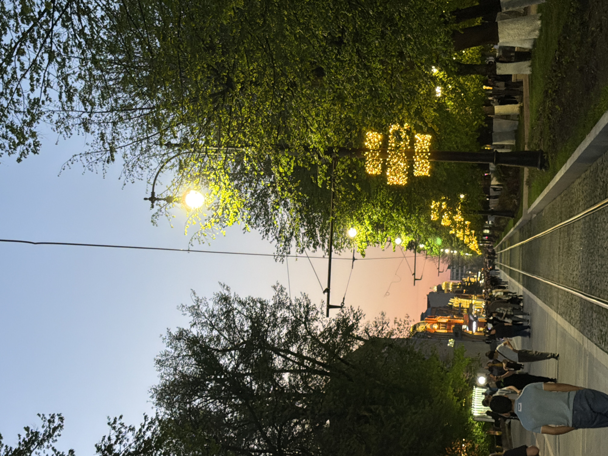 The image shows a street scene in Chermen, Russia, at dusk. A tram line runs down the center of the street, flanked by trees and sidewalks. String lights decorate the trees, and streetlights illuminate the scene. Pedestrians are walking along the sidewalks. The sky is transitioning from daylight to twilight, with a gradient of colors visible. Buildings are visible in the distance, lit up with lights.
