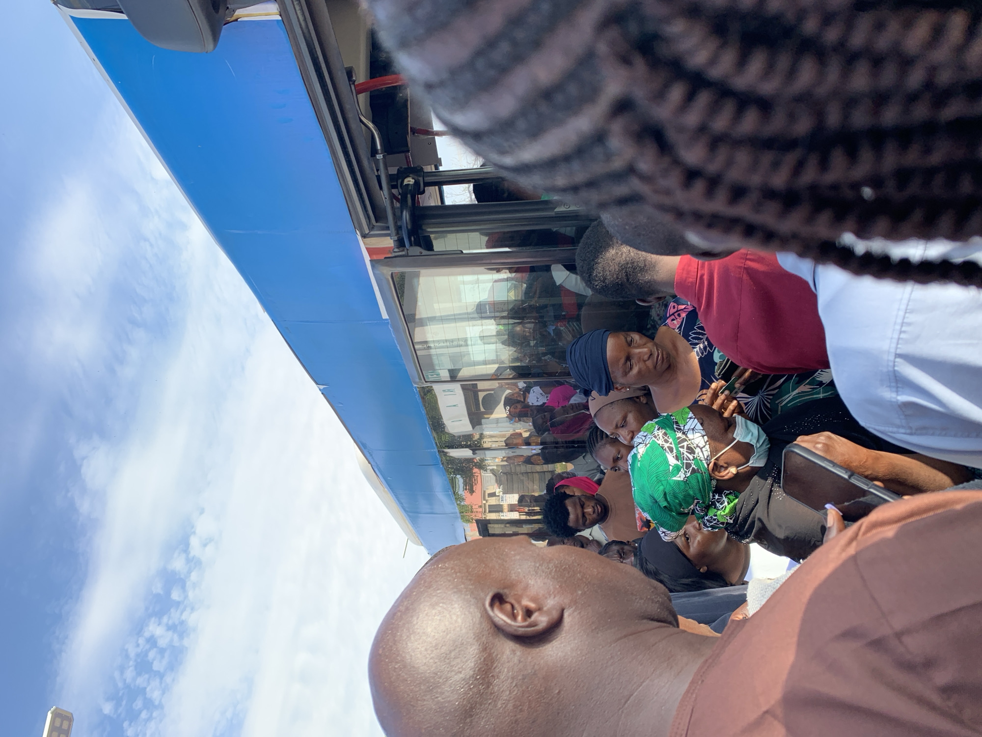 A group of people are gathered on a street in Rafin Par, Nigeria, near a blue bus. The sky is cloudy. The people appear to be boarding or disembarking from the bus. Some are wearing masks. One person is holding a smartphone. The scene is crowded, and the people are close together. The bus is large and appears to be a public transportation vehicle. The overall impression is one of a busy and active urban environment.