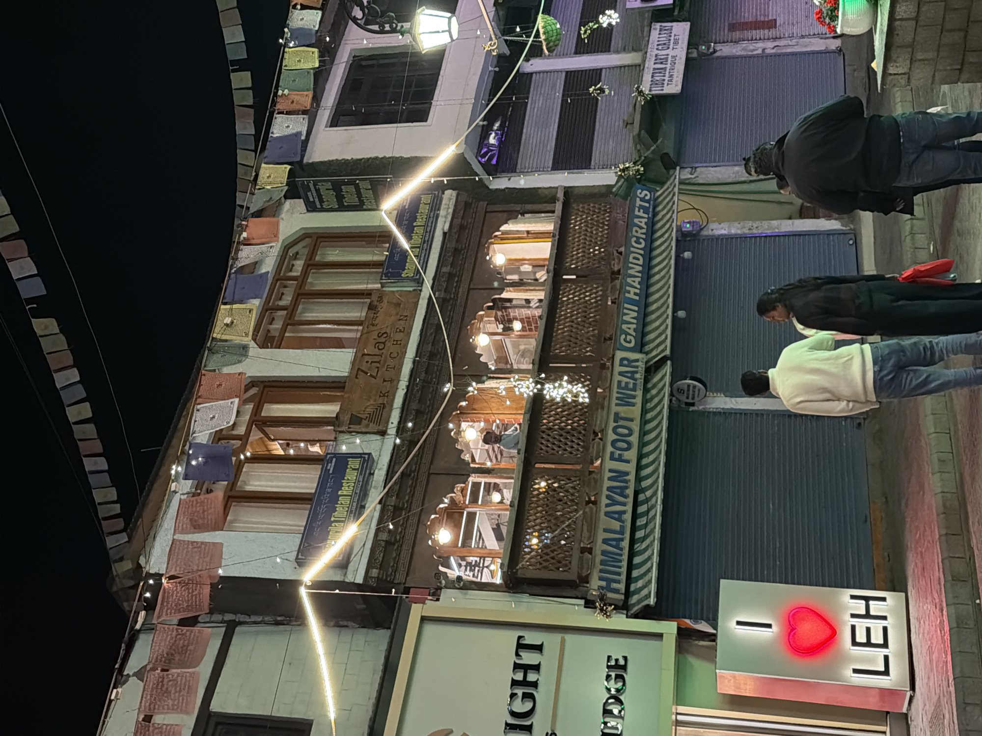 A nighttime street scene in Leh, India features several shops and a large illuminated sign. The sign, prominently displayed on the left, reads "I LOVE LEH" with a glowing red heart. To the right of the sign, a group of three individuals stand on the sidewalk. One person, wearing a light-colored sweater and jeans, faces away from the camera, looking towards the shops. Another person, wearing dark clothing, stands beside them, also facing away. A third person in a black jacket and jeans stands further to the right, also facing away.
The street is lined with shops, including "HIMALAYAN FOOT WEAR" and "GANI HANDICRAFTS." Above these shops, a balcony is visible with several people inside what appears to be a restaurant. Prayer flags are strung across the buildings, and decorative lights illuminate the storefronts and balcony. The overall environment suggests a bustling commercial area in Leh, likely a popular tourist destination.