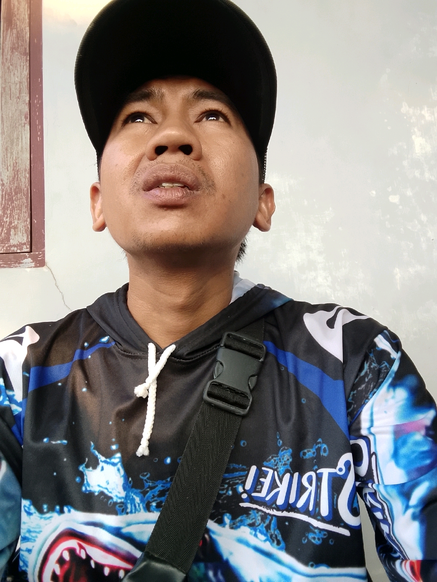 A close-up, waist-up, outdoor selfie shows a person wearing a black baseball cap, looking upwards. The person has short dark hair, dark eyes, and is looking towards the top left of the frame. They have faint stubble on their upper lip and chin, and a small mustache. The person is wearing a black hooded sweatshirt with a vibrant, abstract design featuring a shark's mouth and blue water splashes, and the word "STRIKE!" written in reverse on the right sleeve. A black strap with a black plastic buckle crosses diagonally over the sweatshirt. The background consists of a textured, light-colored wall with some darker imperfections, and a portion of a wooden window frame on the left side of the image. The lighting suggests it is daytime. This scene could be located in Pelaihari, Indonesia, given the person's apparent ethnicity and the typical outdoor setting.
