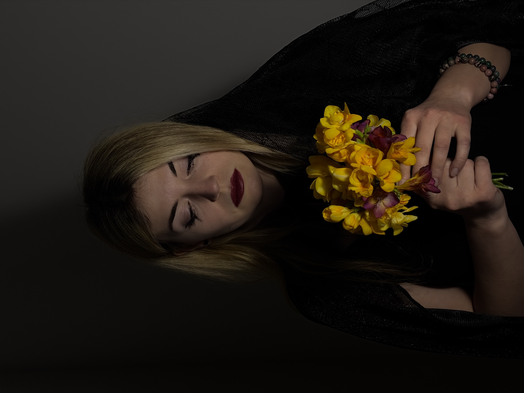 A medium-shot image shows a woman with blonde hair, dressed in black, with her eyes closed. She holds a bouquet of yellow and purple flowers in her hands, which are adorned with a beaded bracelet. The woman is lying down on a dark surface, with a dark background. The scene is dimly lit. There is no indication of a location in Budva, Montenegro, in the image.