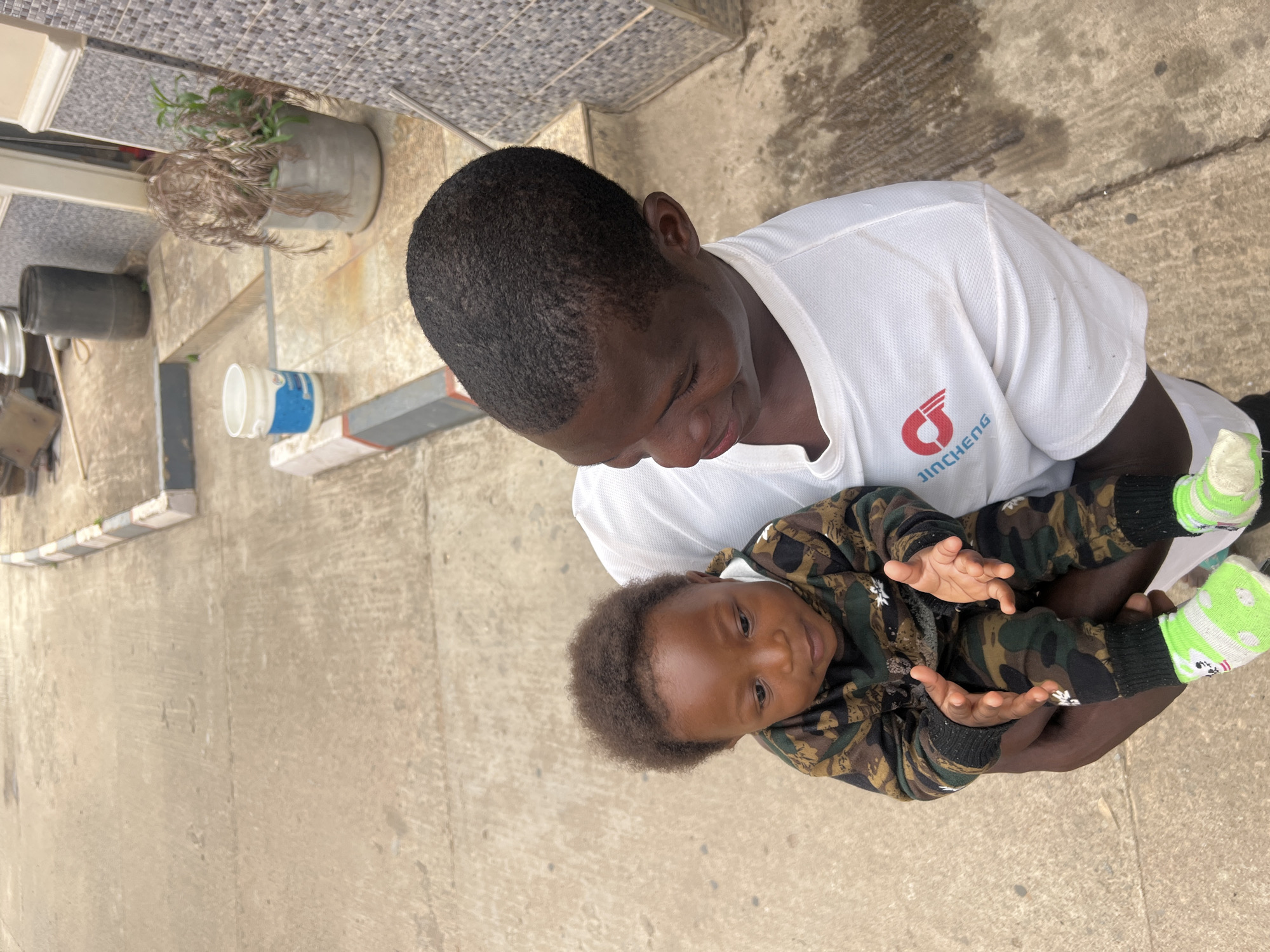 A man in a white T-shirt with a "JINCHENG" logo is holding a baby in a camouflage outfit and green socks. They are outside on a concrete surface. A building with tiles, a plant in a pot, and a white bucket are visible in the background. The location is likely Jos, Nigeria, based on the context provided.