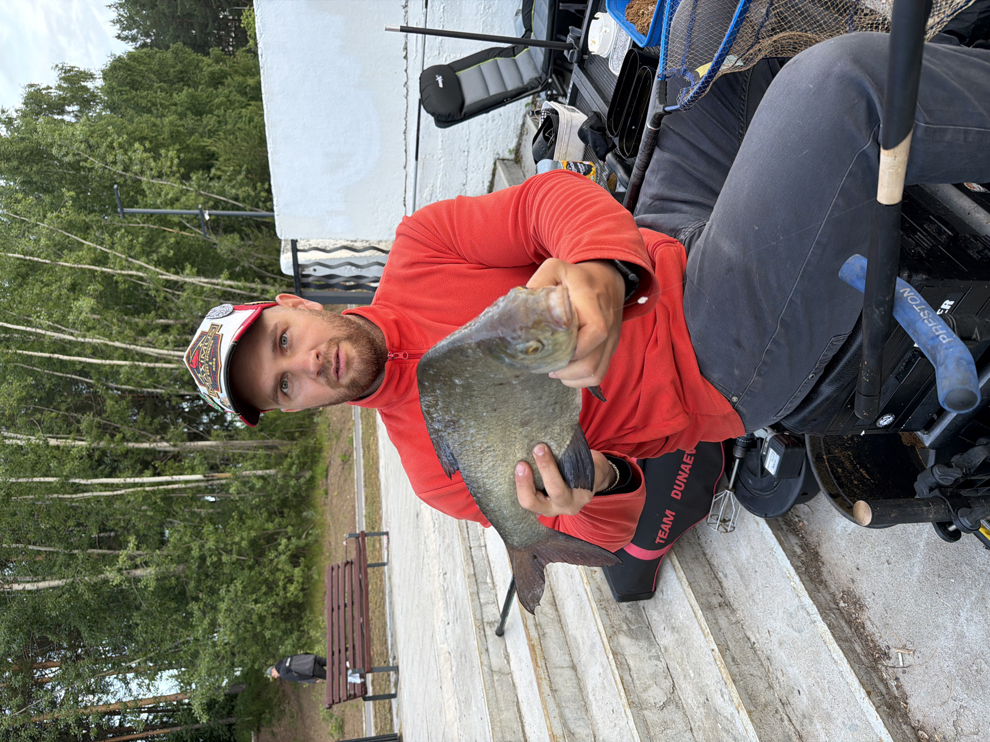 A man, likely a fisherman, is the central subject of this outdoor portrait, positioned from the waist up and looking directly at the camera. He is wearing a red long-sleeved fleece top, dark pants, and a white and red baseball cap. He has a light beard. The man is proudly holding a large, greyish-green fish, possibly a bream or carp, in both hands.
The setting appears to be a fishing spot, possibly along a riverbank or lake in Chaikovsky, Russia, characterized by concrete steps or a paved platform where the man is seated or kneeling. Behind him, various fishing equipment is visible, including a black fishing seat or chair, a blue fishing net, a fishing rod, and several containers, one appearing to hold bait or groundbait. A blue hose or pipe-like object with the word "PRESTON" printed on it is also near his leg. On his left sleeve, the text "TEAM DUNAEV" is clearly visible in white letters. His baseball cap has some partially legible text, possibly "MAXIMUS".
In the background, there's a lush green tree line, with some slender birch trees and dense foliage suggesting a natural, park-like environment. A white building or structure is partially visible behind the trees on the right side. Further in the distance, a brown wooden park bench is present, and a solitary figure is partially visible walking near it. The sky is overcast, indicating daytime with soft, diffused lighting. The overall scene captures a moment of a successful fishing outing.