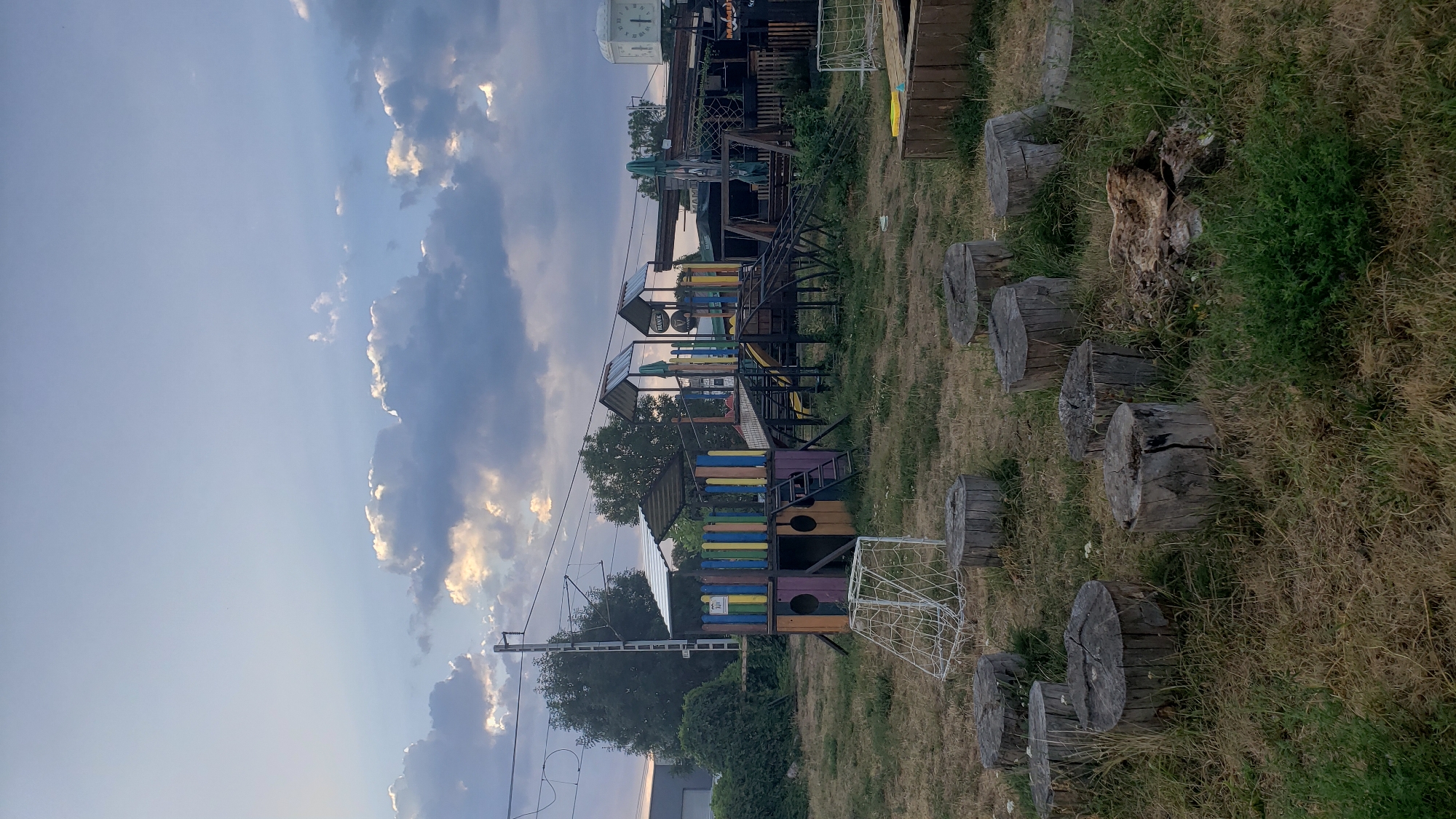 This image captures an outdoor play area, likely situated in the city of Prešov, Slovakia. The scene is dominated by a colorful, multi-level playground structure with slides, climbing elements, and a small wooden fort at its base, painted in vibrant hues of blue, yellow, green, and purple. In the foreground, several tree stumps are arranged in a circular pattern on dry, sparse grass, serving as natural seating or stepping stones. A white, net-like structure, possibly a small goal, is positioned near the stumps. In the background, a utility pole with overhead wires suggests proximity to a railway line or power infrastructure. The sky above is a pale blue, partially obscured by scattered, fluffy clouds, some of which are illuminated by the setting sun, casting a warm glow. The overall environment appears somewhat unkempt, with overgrown grass and a general sense of rustic charm. No people are visible in the frame.