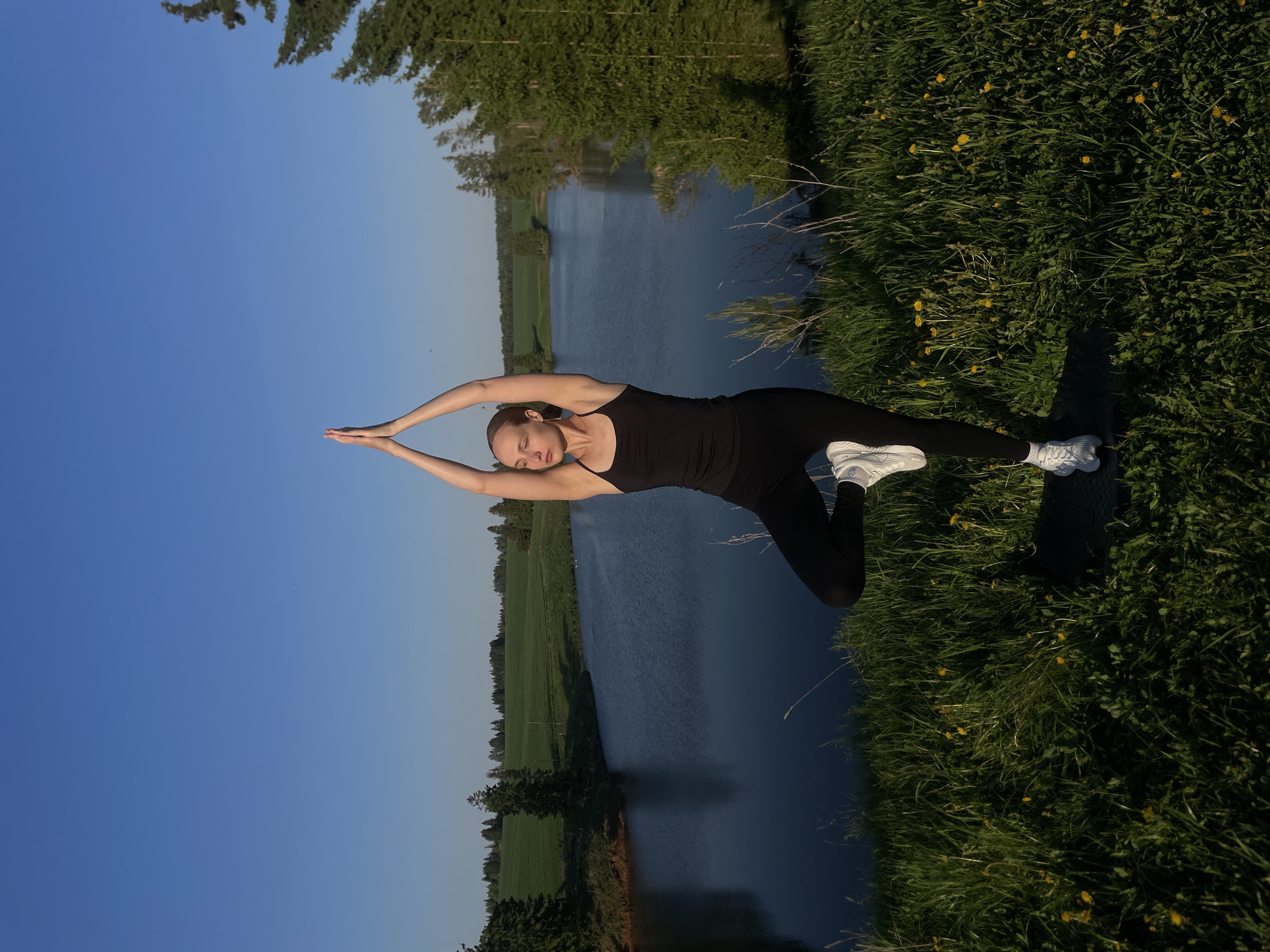 A woman in black athletic wear and white athletic shoes is performing a yoga pose with arms extended above her head on a grassy bank. A body of water lies behind her, with a grassy field and trees on the opposite bank. The sky is blue. The photograph was taken in Podshivalovo, Russia.