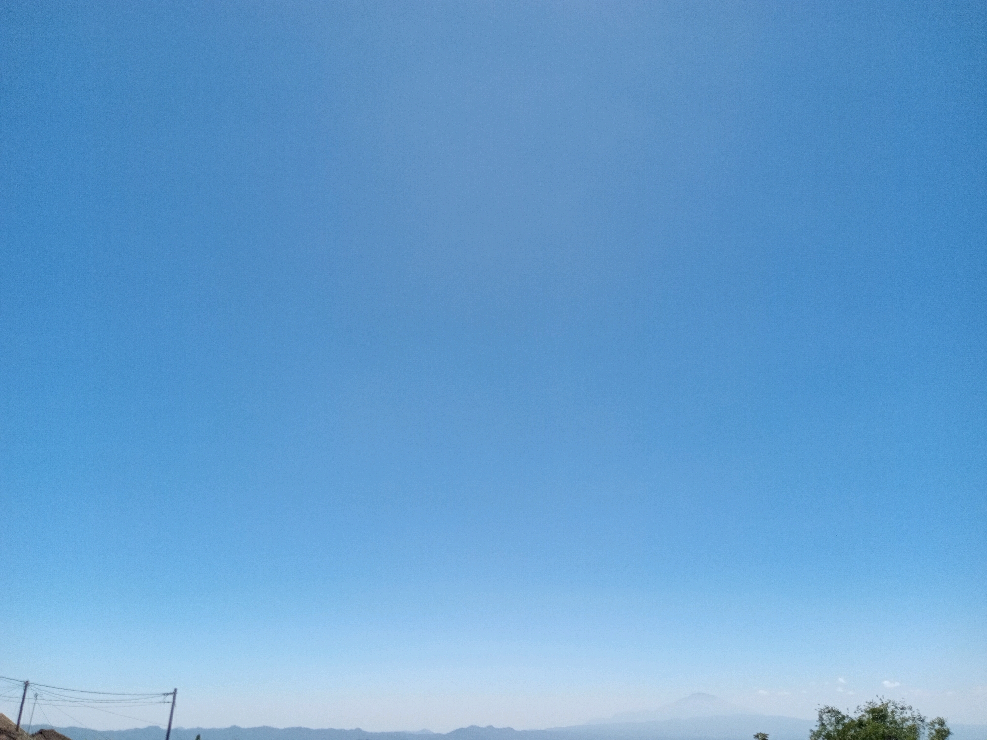 The image captures a clear, bright blue sky above a distant, hazy landscape. The horizon is marked by a range of mountains or hills. In the foreground, on the left side of the image, there are utility poles with wires. On the right side of the foreground, a dense cluster of green trees is visible. The overall scene suggests an outdoor environment in a somewhat rural or suburban setting. The location is identified as Gandurejo, Indonesia.