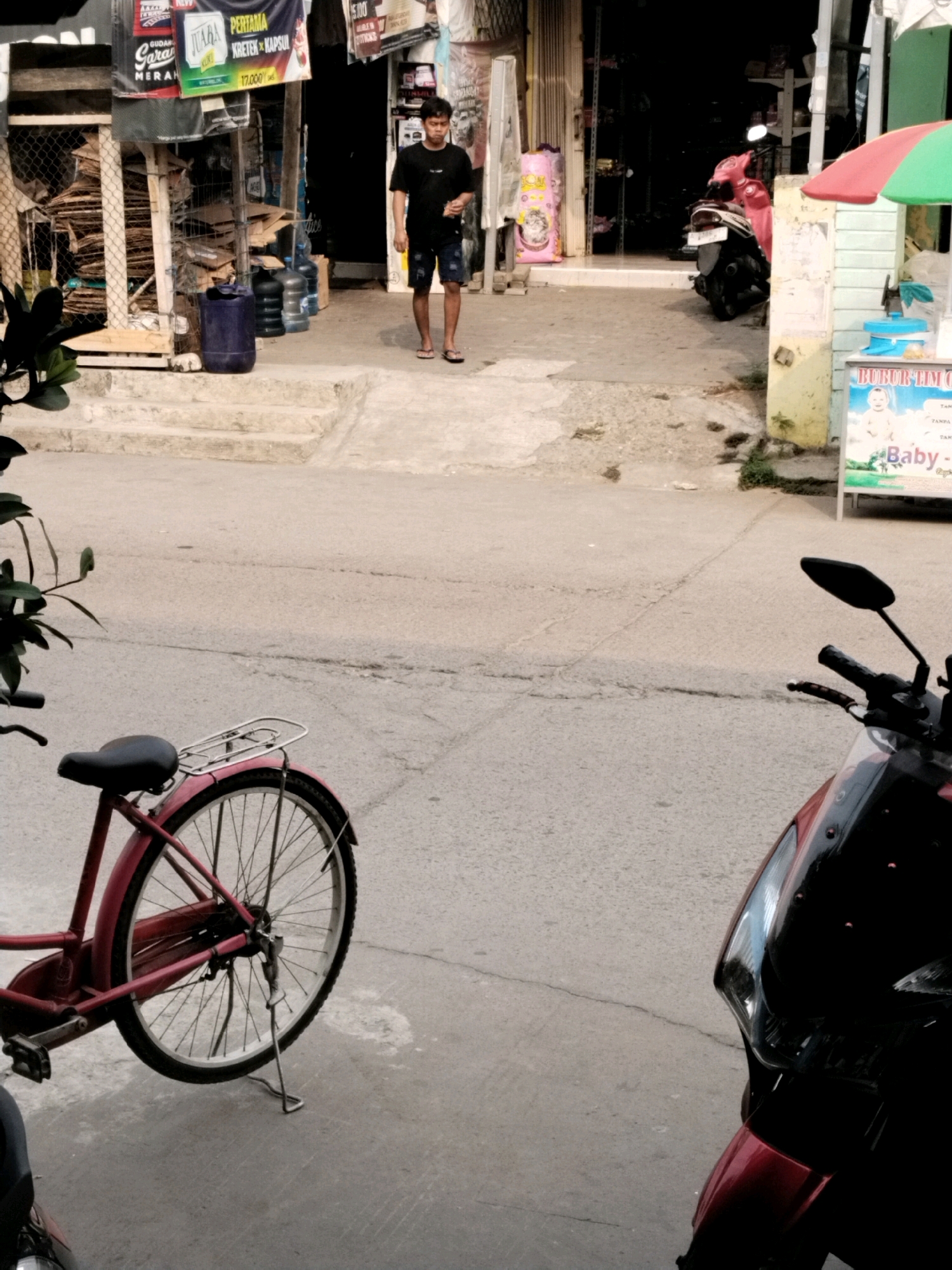 The image depicts a street scene in Serang, Indonesia. In the foreground, a red bicycle is parked, with its rear wheel and kickstand visible. To the right, the front of a dark-colored motorcycle, likely red and black, is visible, including its handlebars and mirrors. In the middle ground, a person wearing a black t-shirt and shorts walks away from the camera. Behind the person is a storefront with various items displayed, including what appears to be bottled water and cardboard boxes. A pink bag with a cartoon design is also visible near the storefront entrance. A red and green umbrella is situated to the right of the storefront. The ground is paved with asphalt, showing some cracks.