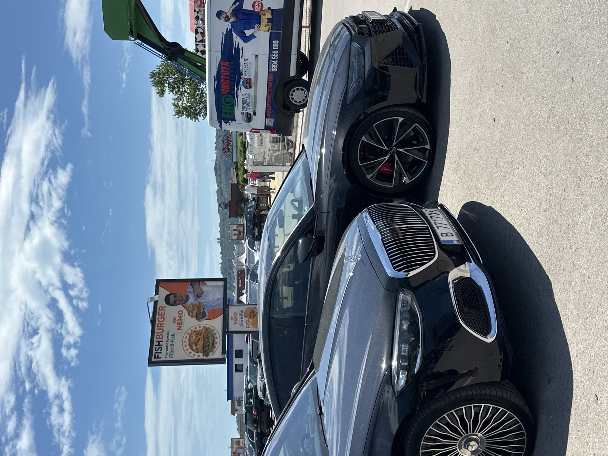 The image is a low-angle, medium-shot taken outdoors on a sunny day, primarily featuring two black cars parked next to each other. The front of a black Mercedes, showing its grill, headlight, license plate (B 7717XK), and front tire, is in the foreground. Adjacent to it is an Audi. In the background, there is a commercial vehicle for "Eko Chistota" (an environmental cleaning service), with text in Bulgarian. The location appears to be in or near Varna, Bulgaria, evidenced by Bulgarian text on the commercial vehicle. A tall green amusement ride tower is behind the commercial vehicle. A billboard advertising "Fish Burger Nemo" with images of fish burgers is visible. Other parked cars are further in the background. The sky is blue with scattered clouds.