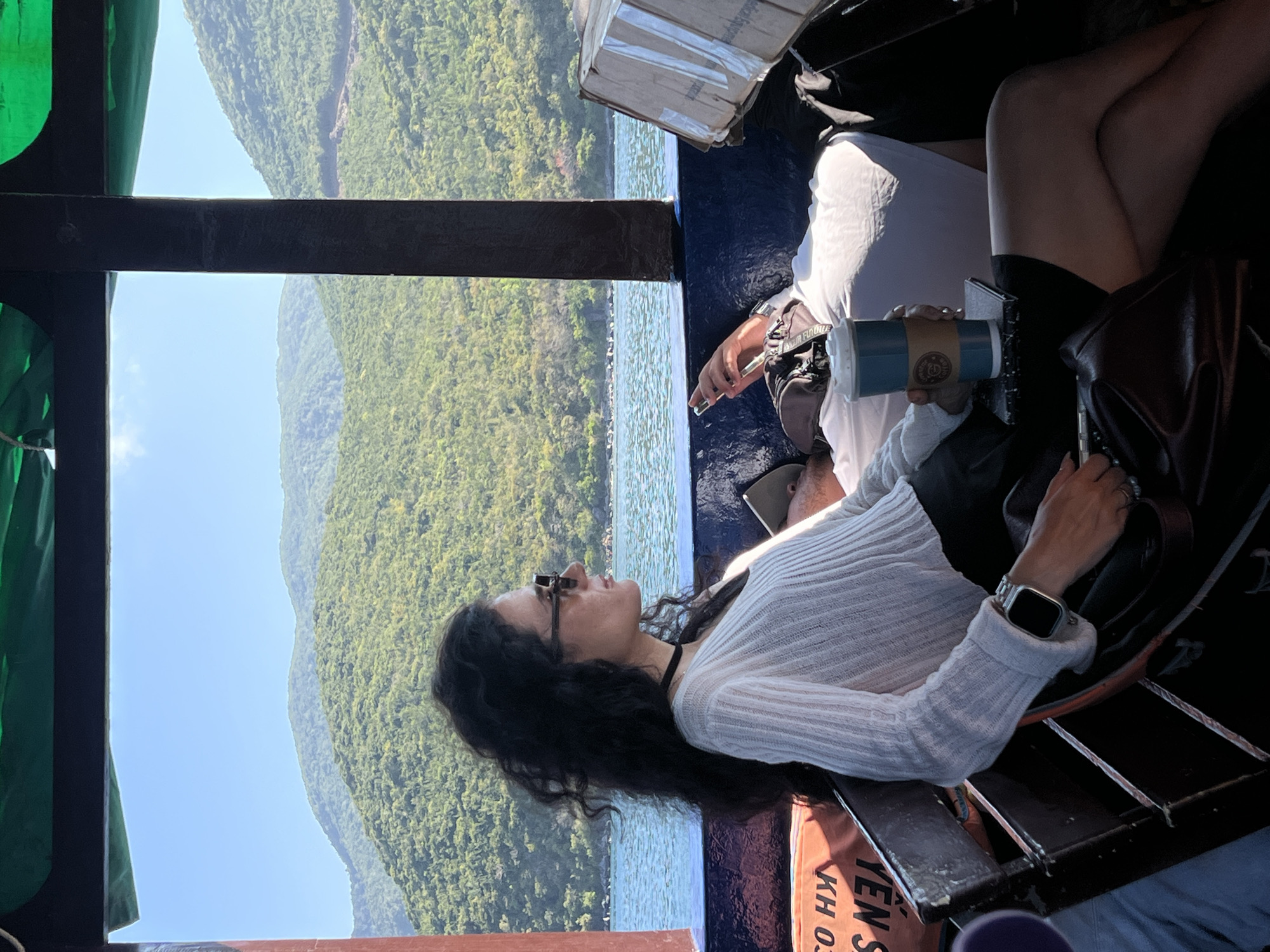 A young woman with dark hair and sunglasses is reclining on a boat, her head tilted back and her face turned towards the lush, green mountains visible across the water. She is wearing a white sweater and an Apple Watch. The boat is moving across the water, offering a scenic view of the mountains and sky. Another person on the boat holds a coffee cup. The location is Nha Trang Ward, Vietnam.