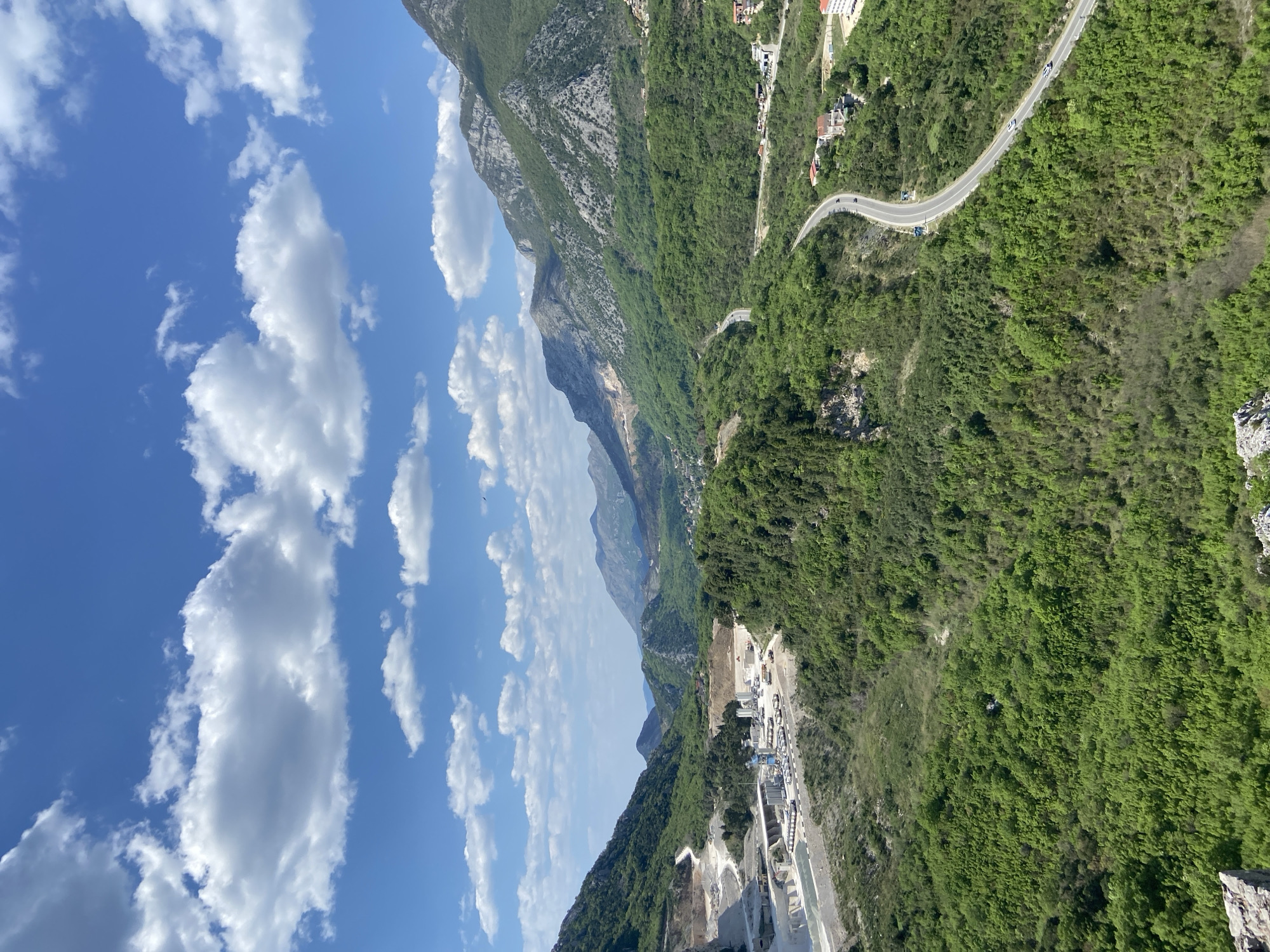 The image shows a landscape in Zagrađe, Montenegro. The sky is blue with scattered white clouds. Mountains covered in dense green vegetation dominate the view. A winding road with a few visible vehicles snakes through the mountains. In the foreground, there is what appears to be an industrial area with white structures and machinery, possibly a quarry or a processing plant.