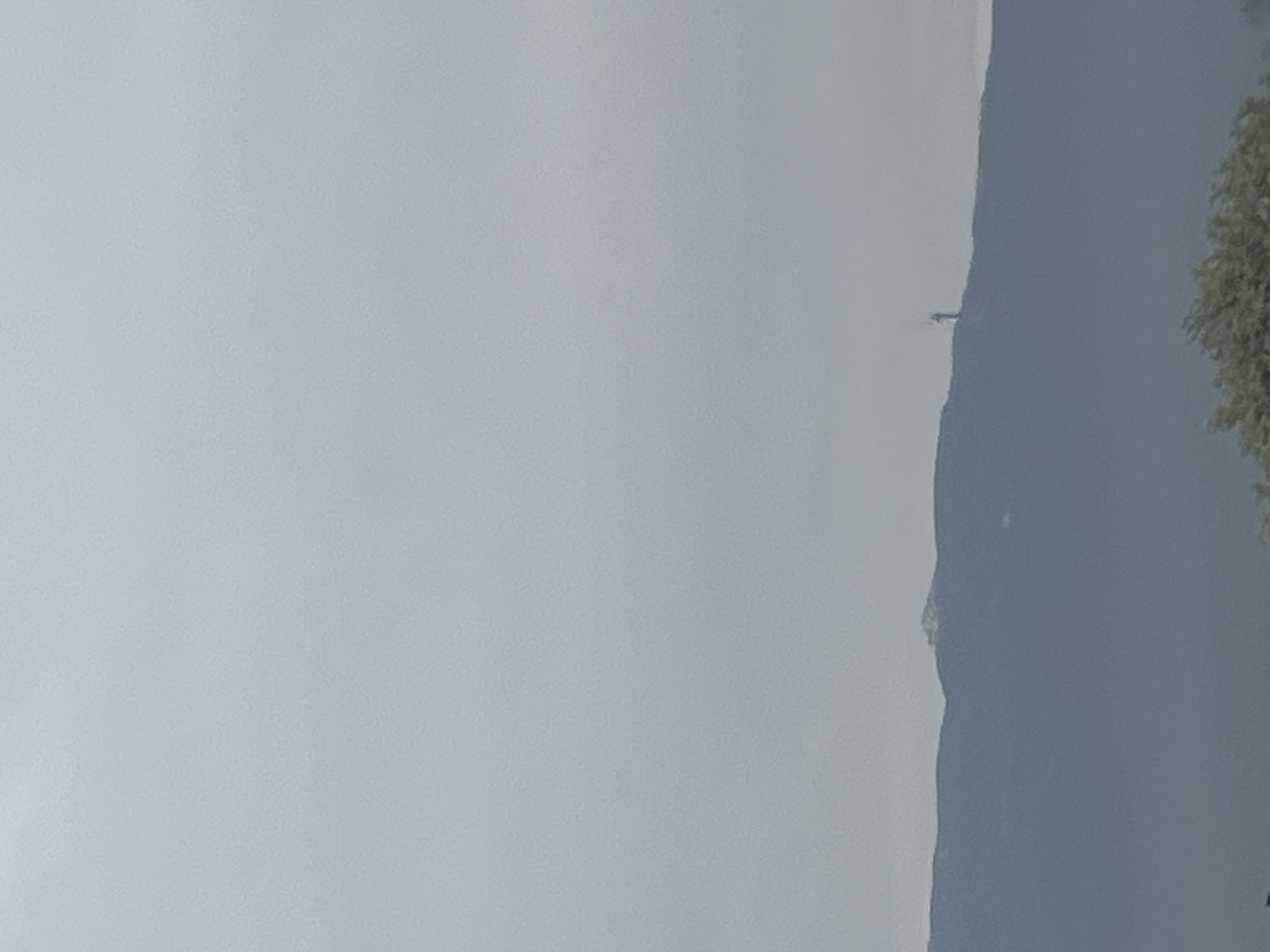 A landscape scene features a mountain range obscured by a hazy, grey sky. A section of the mountain range on the right side is slightly more visible, with some trees or foliage clinging to its slopes. In the center of the shot, close to the mountaintop, there's a faint outline of a tower or structure. The overall lighting and atmospheric conditions render distant details obscure. The location is Unknown, Unknown.