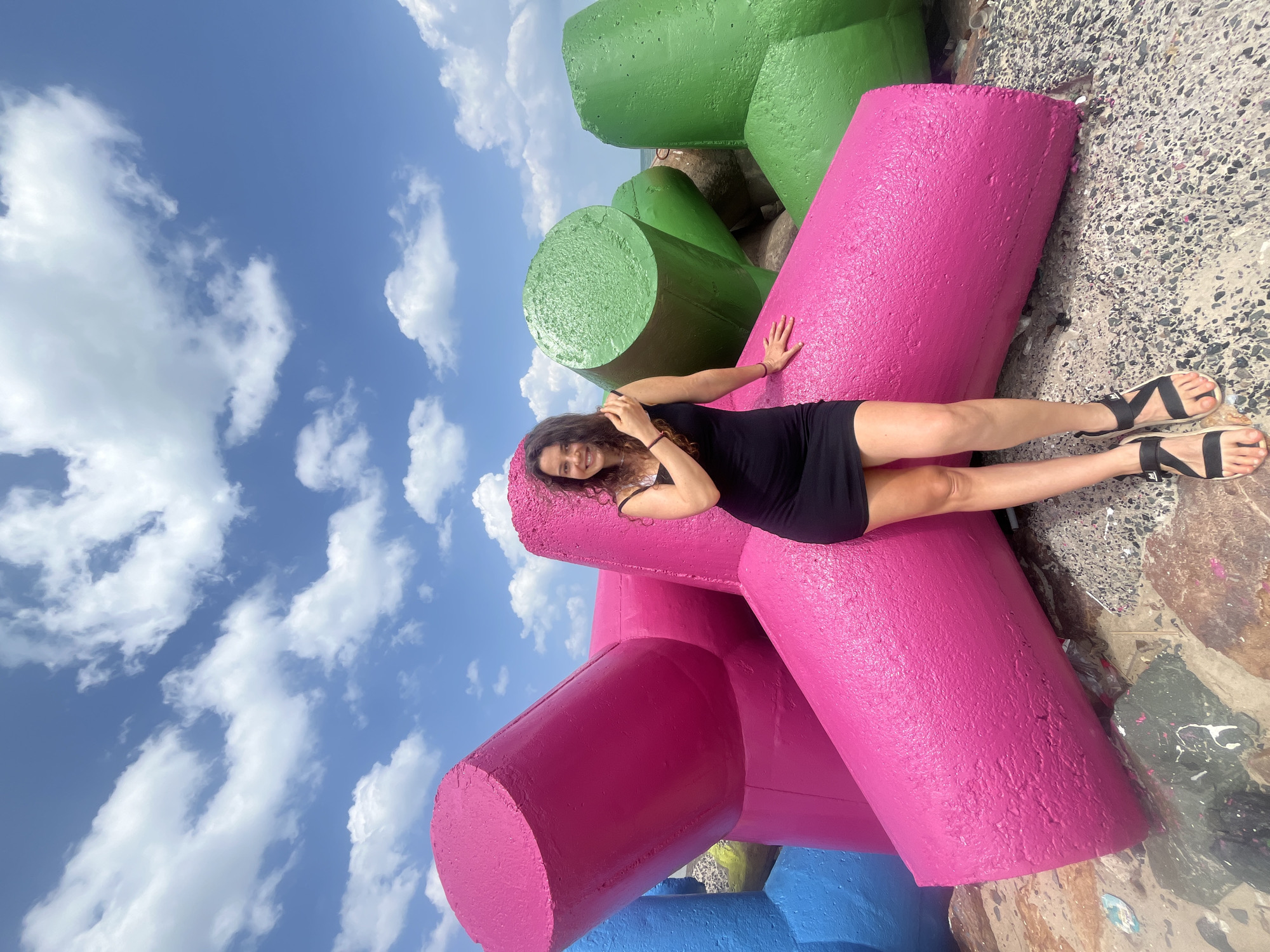 A young woman stands on a rocky surface, leaning against large, brightly painted concrete structures. The woman is wearing a black dress and black sandals. The concrete structures are tetrapods, a type of coastal defense structure, painted in vibrant pink, green, and blue. Behind the tetrapods, the sky is blue with scattered white clouds. The scene appears to be outdoors, likely near a body of water, given the tetrapod structures. The location is not specified.