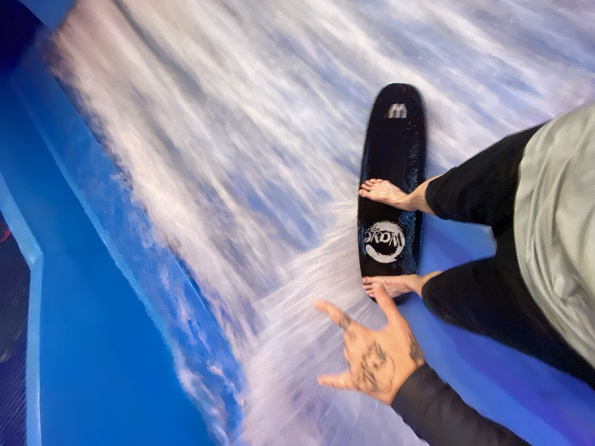 The image captures a first-person perspective of someone standing on a surfboard in a wave pool. The person's legs and feet are visible, along with a tattooed hand in the upper left corner, making a "rock on" hand gesture. The surfboard is black and has a circular logo with the word "Wave" on it. The environment is a bright blue wave pool with white water effects. The scene suggests an indoor water park or similar recreational facility. There is no information in the image that places this scene in Khabarovsk, Russia.