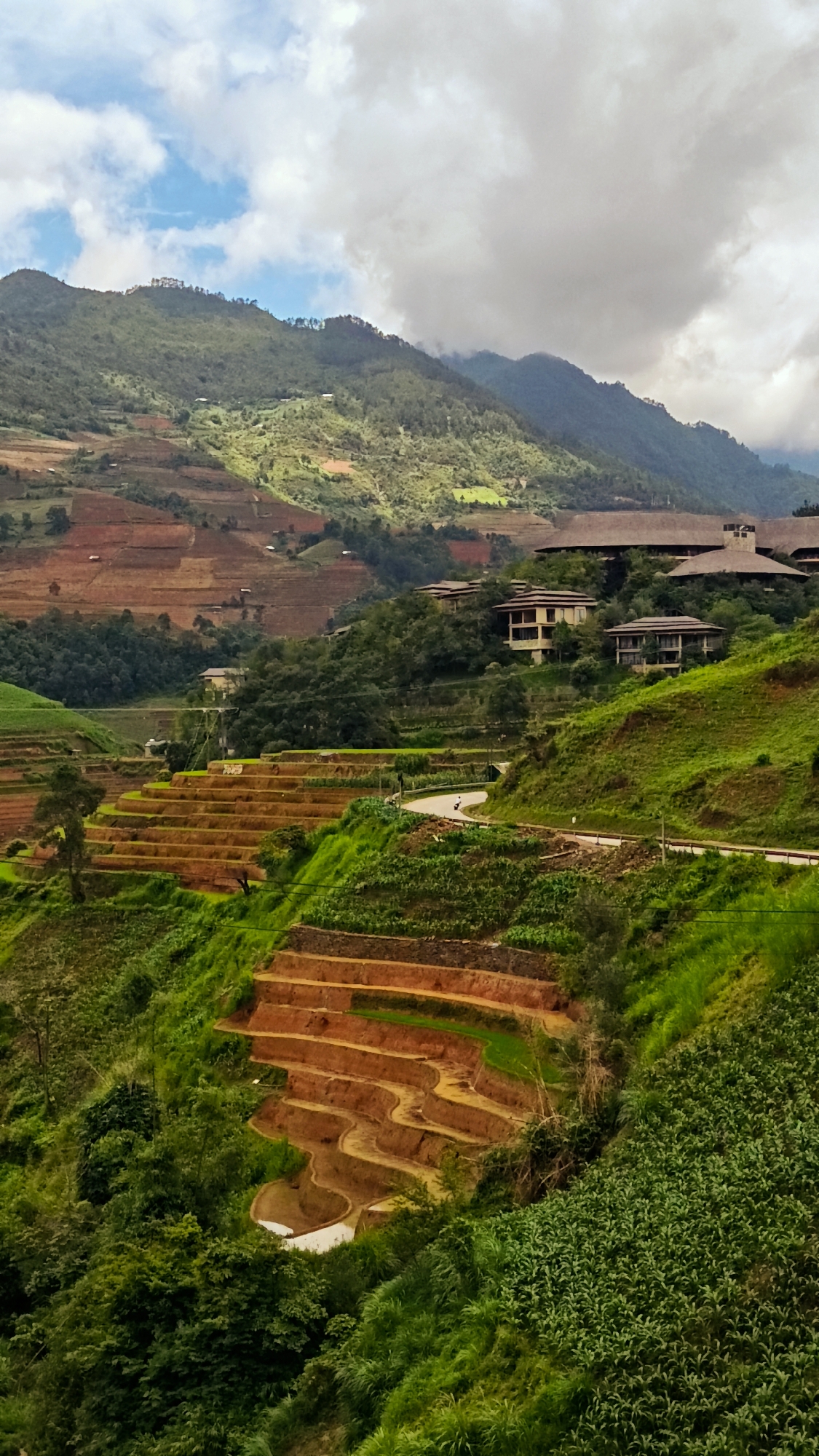 This vertical photograph captures a mountainous, terraced landscape in Mù Cang Chải, Vietnam. The sky in the upper frame is partly cloudy, displaying patches of bright blue on the left and dense grey clouds obscuring the upper right portions and distant mountain peaks. The background is dominated by large, green mountains covered in dense foliage. Numerous rice terraces, cut into the hillsides, are visible throughout the mid-ground and foreground. These terraces display varied states; some are reddish-brown, indicative of freshly tilled earth, while others are vibrant green with growing crops. Lower terraces in the foreground appear partially flooded with water. Several buildings are situated on the slopes; a cluster of large structures with dark, possibly thatched roofs is prominent on a ridge in the upper right, with additional smaller, multi-story residences further down. A paved, winding road traverses the mid-ground, extending from the right towards the center. Lush green vegetation, including trees and dense groundcover, is abundant across the uncultivated areas. A small, dark animal is visible on the road in the mid-right. No human figures are clearly discernible.