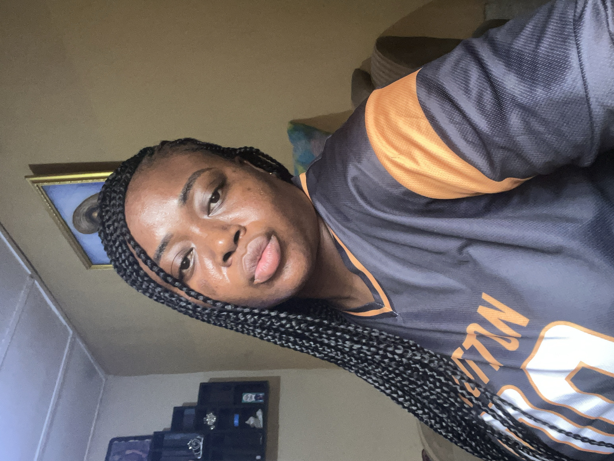 A person with braided hair, wearing a black jersey with orange accents, is oriented on a vertical axis. The setting appears to be indoors, possibly in Madalla, Nigeria, given the user's location. Behind the person, there is a wall, and in the background, there's a television and some electronics. A framed picture hangs on the wall.