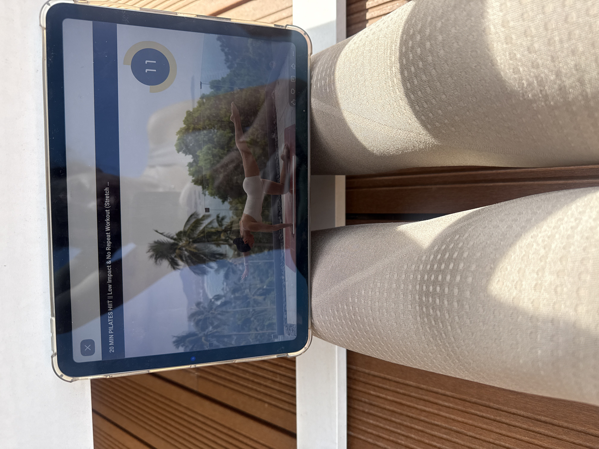An overhead shot captures a tablet, likely an iPad in a clear protective case, positioned on a slatted wooden surface, which appears to be outdoor decking. To the right, a section of light-colored, textured outdoor furniture or cushioning is visible. The scene is brightly lit by natural sunlight, casting subtle shadows and indicating a clear, sunny day.
The tablet screen displays a workout video, showing a woman in a light-colored leotard performing a side-plank-like pose with one leg raised, against a backdrop of palm trees and a body of water, suggesting a tropical or exotic location. On-screen text from the video reads "20 MIN PILATES HIIT || Low Impact & No Repeat Workout (Stretch ...)", with a circular progress indicator showing the number "11". A "X" icon, likely a close button, is also visible in the top left corner of the video player interface.
The image portrays an individual engaging in a fitness activity, watching an exercise video outdoors, perhaps on a patio or balcony. The bright weather, outdoor furniture, and wooden deck suggest a leisure setting, possibly during a vacation or relaxing day in Vila Nova de Gaia, Portugal.
