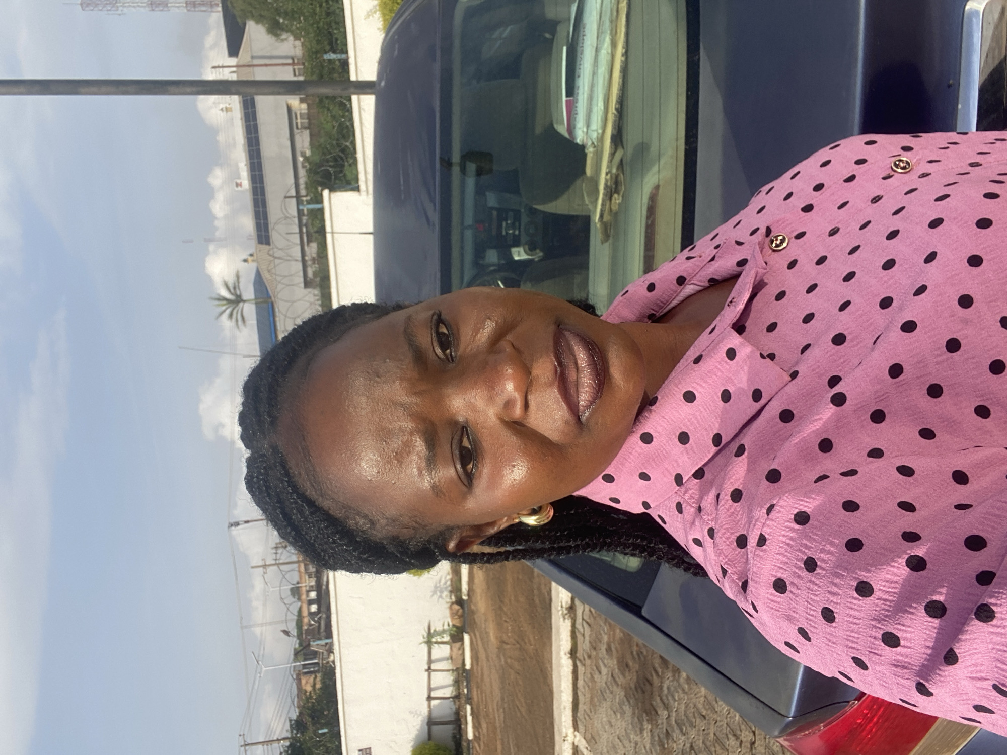 The image shows a woman standing next to a dark blue car. She is wearing a pink polka dot shirt and gold hoop earrings. Her hair is braided. The scene appears to be outdoors, with a cloudy sky and some buildings in the background. There is a palm tree visible. The context provided indicates the location is Bukuru, Nigeria. The woman is looking towards the camera with a slight smile. The time of day appears to be daytime, likely morning or afternoon, given the lighting. The weather seems overcast. There is no visible text in the image.