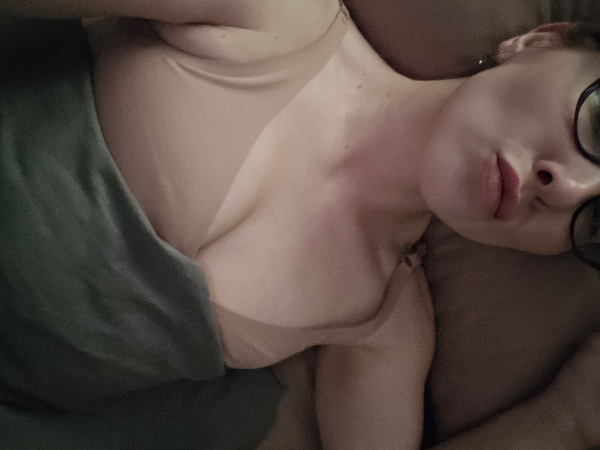 A woman with glasses is lying on a pillow, covered with a gray blanket. She is wearing a beige bra. Her face is turned towards the camera, and she appears to be resting. The location is unknown.
