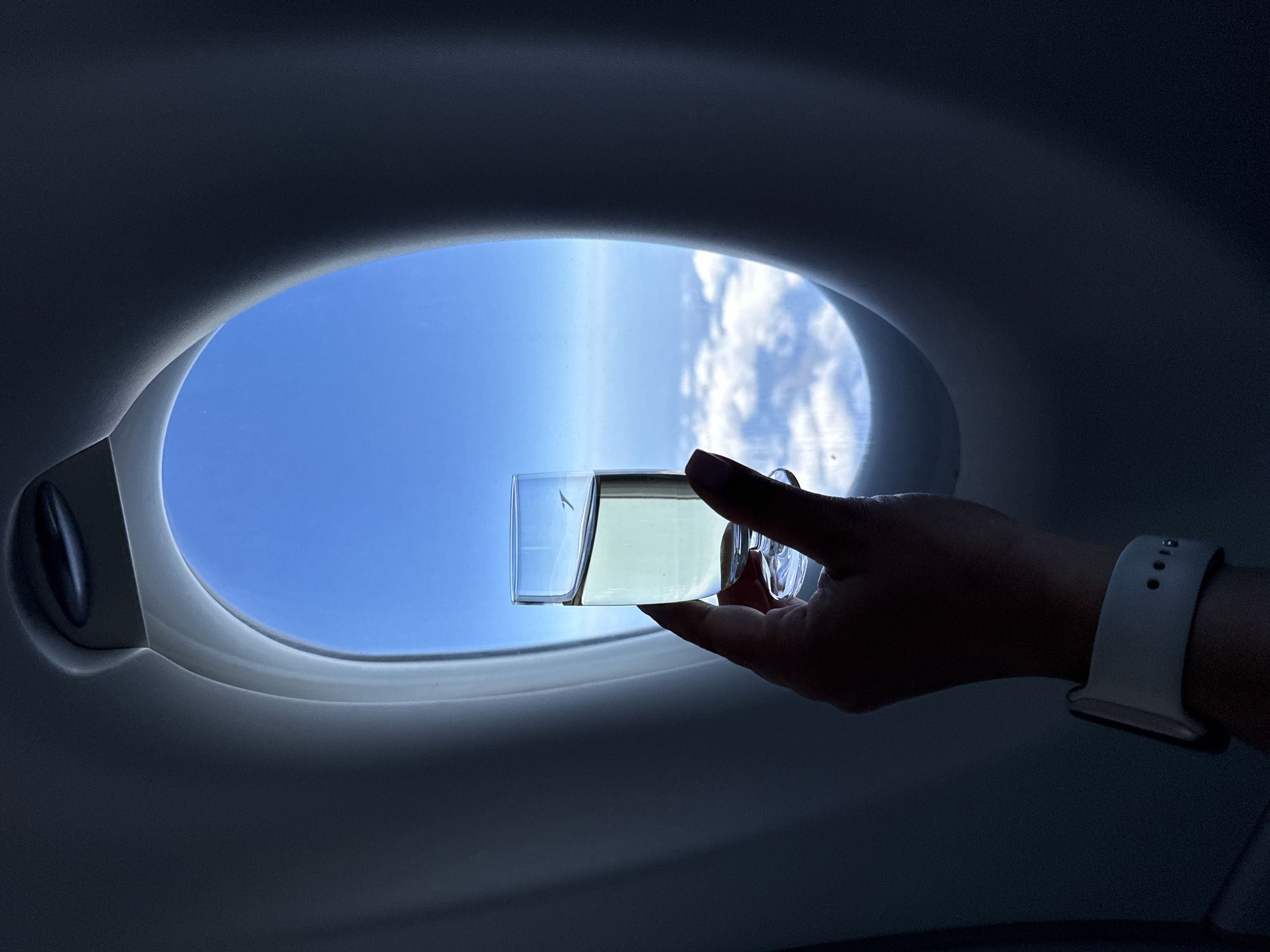 The image shows a hand holding a glass of clear liquid with a white smartwatch on the wrist, in front of an airplane window. Outside the window, there is a clear blue sky with some white clouds below. The interior of the airplane cabin is dark and out of focus, framing the window. There is no discernible indication of the city of Rudeni, Romania.