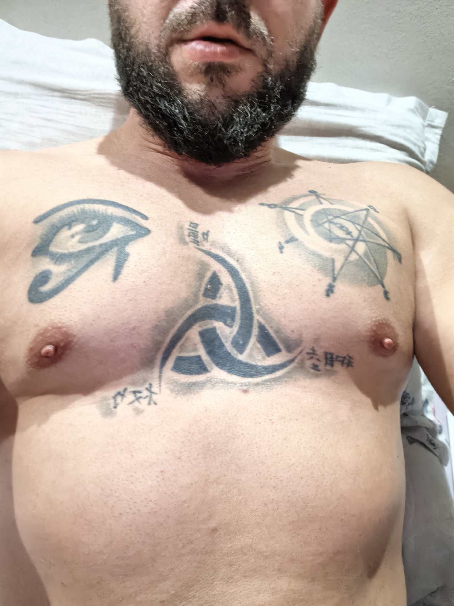 A bearded man is lying on a bed, with his upper torso visible. He has several tattoos on his chest, including an eye, a Celtic knot, a design with stars and circles, and some Asian characters. The background shows a pillow and bedsheets. The location is unknown.