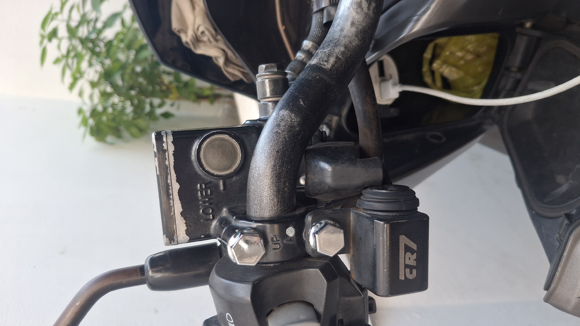 The image shows a close-up view of the handlebars of a motorcycle. The handlebars are black and appear to be slightly worn. Attached to the handlebars are various controls, including a brake reservoir with "LOWER" visible on it, a lever, and a small black accessory with "CR7" etched onto it. A white cable is also visible, plugged into a port located in an open compartment beneath the handlebars. The background is blurred, but suggests an outdoor setting with a white wall and greenery. The image is taken from a slightly high angle, looking down at the handlebars. There is no indication of the specific location within Tasikmalaya, Indonesia, beyond the general context of a motorcycle handlebar.