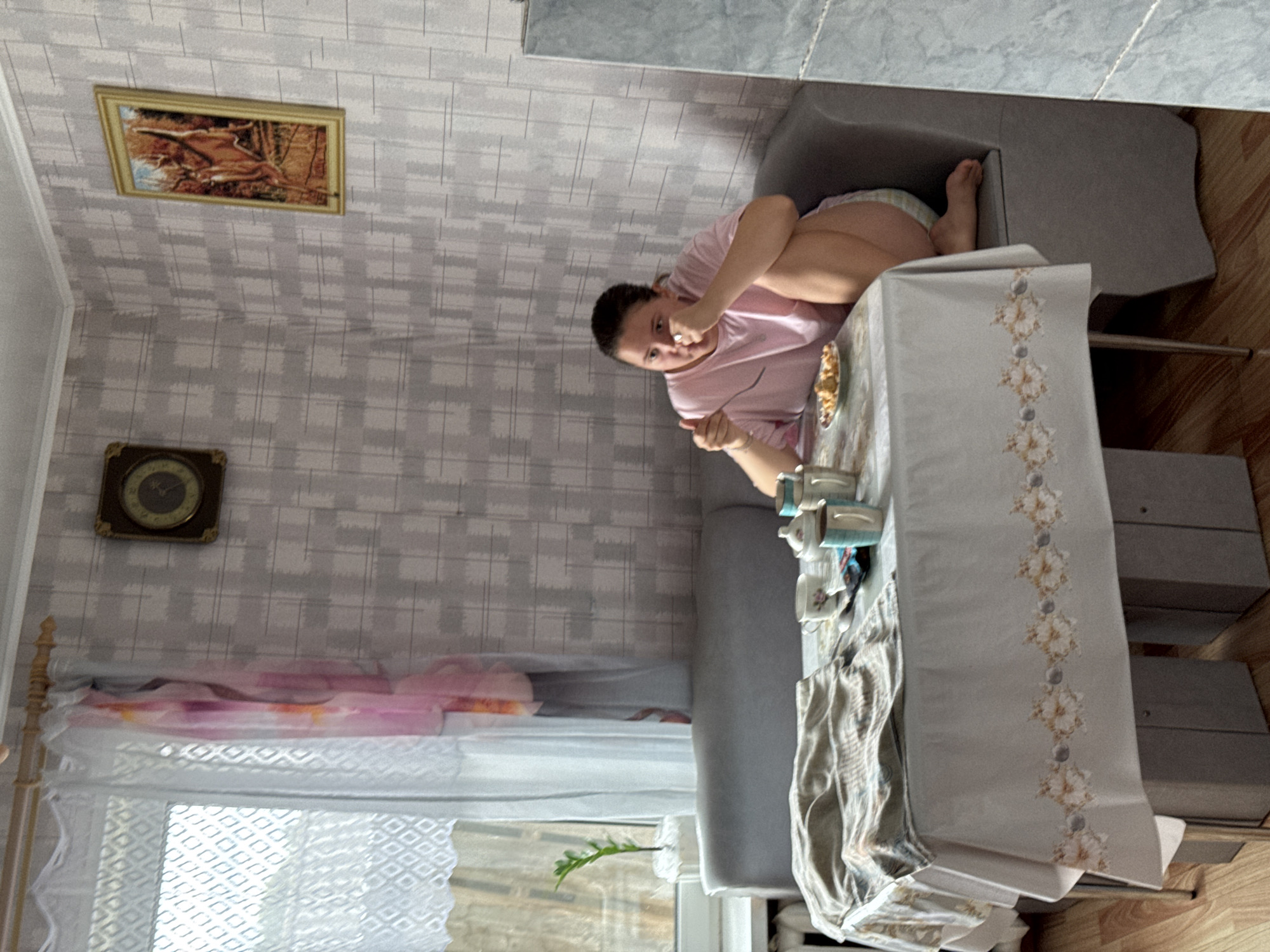 A young woman with dark hair pulled back is seated at a table in a room, wearing a pink t-shirt and shorts. She is holding a fork in her right hand and bringing it towards her mouth. On the table in front of her is a small plate with food, along with several cups and a sugar bowl. A tablecloth with a floral pattern covers the table, and a plant sits on a windowsill to the left. The walls are covered with patterned wallpaper. A clock hangs on one wall, and a framed picture of a deer is on another. The woman appears to be eating a meal in a domestic setting, likely in a home in the village of Borodulino, Russia, given the context. The lighting suggests it is daytime.