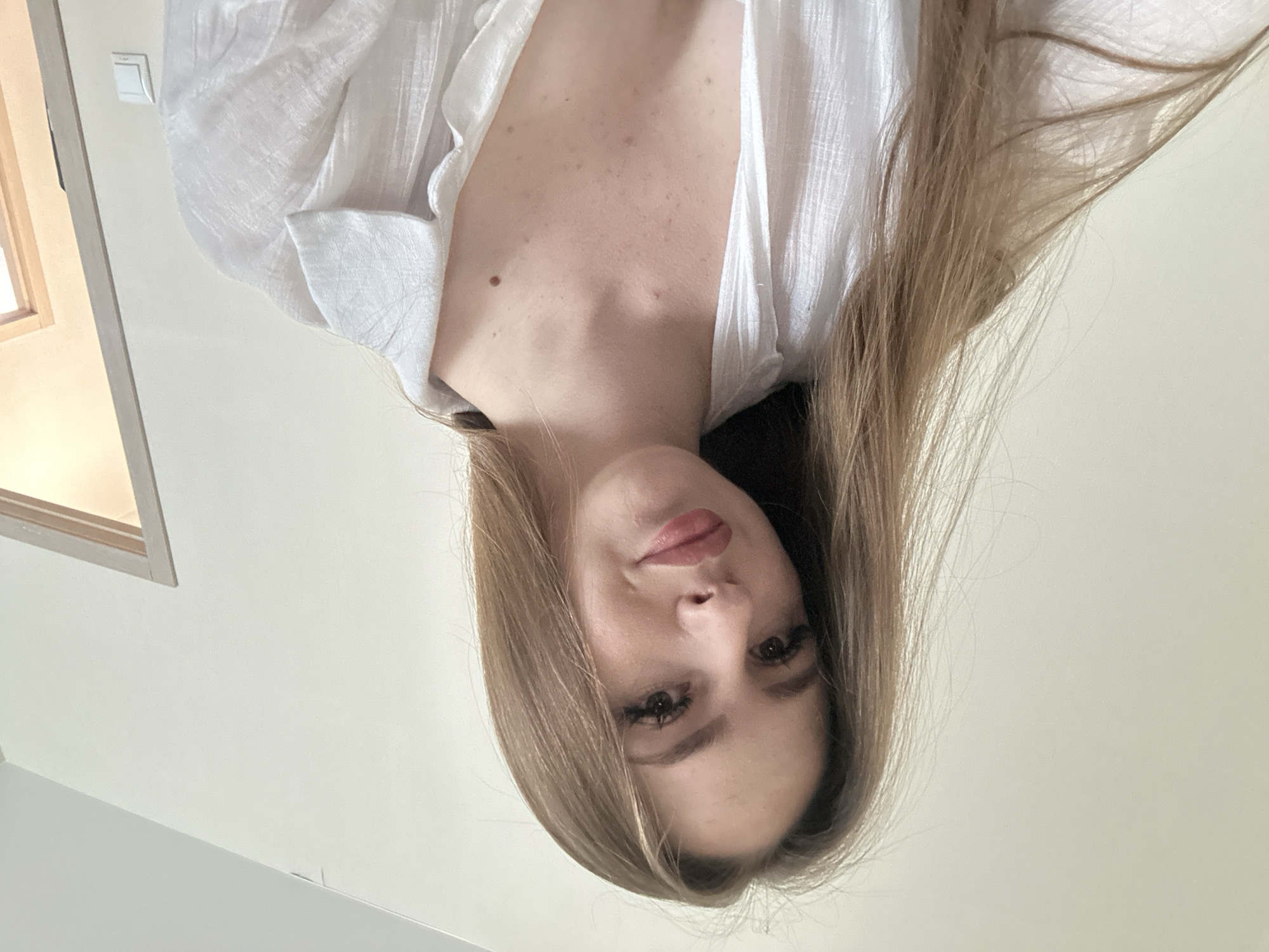 A young woman with long, light brown hair and fair skin is depicted in a selfie. She is wearing a white, loose-fitting shirt that is unbuttoned, revealing her décolletage. She has noticeable eyelashes and light makeup on her lips and eyes. The background is a plain, light-colored wall, with a doorframe visible to the right of the frame. There are a few small marks or blemishes on her chest. The lighting is soft and natural, suggesting an indoor setting.