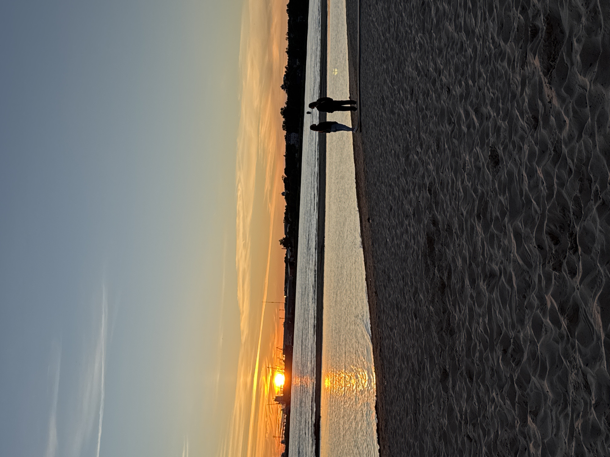 This is a vertical, eye-level, outdoor shot of a beach at sunset, likely taken in Tallinn, Estonia, near the port. Two individuals are silhouetted against the twilight sky as they stand on the sand near the water's edge, facing the setting sun. The sun, a brilliant orange orb, is low on the horizon, casting a shimmering reflection on the calm water. In the distance, a cityscape with cranes and buildings is visible, suggesting the presence of a port or industrial area. The foreground is dominated by textured sand dunes, characteristic of a beach environment. The sky transitions from a pale blue at the top to warm hues of orange and yellow near the horizon, indicative of a sunset.
