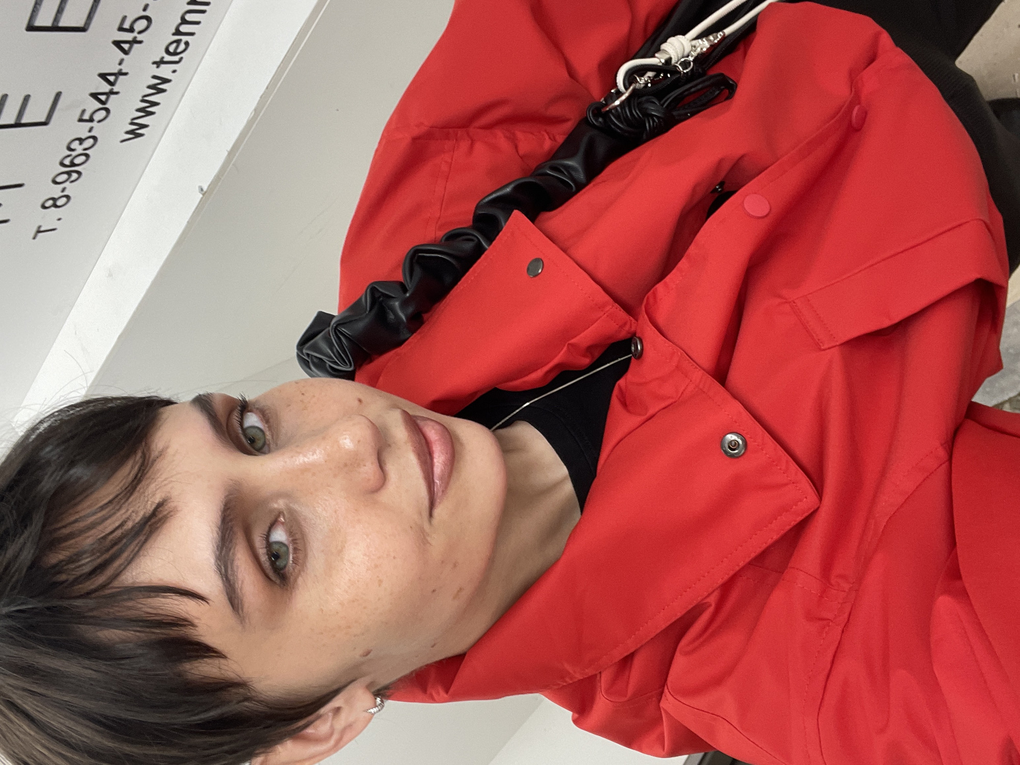 The image is a close-up, slightly angled shot of a person wearing a bright red jacket. The person has short, dark hair with bangs, light skin with freckles, and light green eyes. They are wearing an earring on their left ear and a black shirt under the jacket. Part of a white shelf or counter is visible on the left side of the frame, displaying printed text that includes the phone number "T. 8-963-544-45-" and the website "www.tem". Behind the person, and draped over the jacket, there appears to be a black scrunchie and some type of lanyard or cord. There is no information indicating the location of the photo being Izhevsk, Russia.