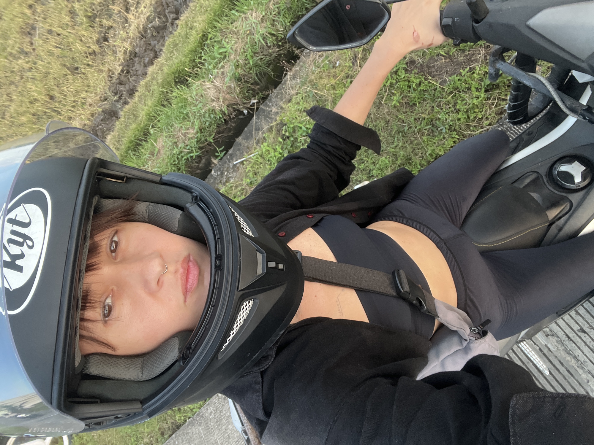 A woman is seen in a close-up, first-person shot while sitting on a black and white scooter. She is wearing a black KYT helmet, black leggings, a black sports bra, and a black button-down shirt. The helmet is fastened, visible by the strap across her chest. She has a nose ring and is looking directly at the camera. The background shows a grassy, ditch-like area, possibly in North Kuta, Indonesia.