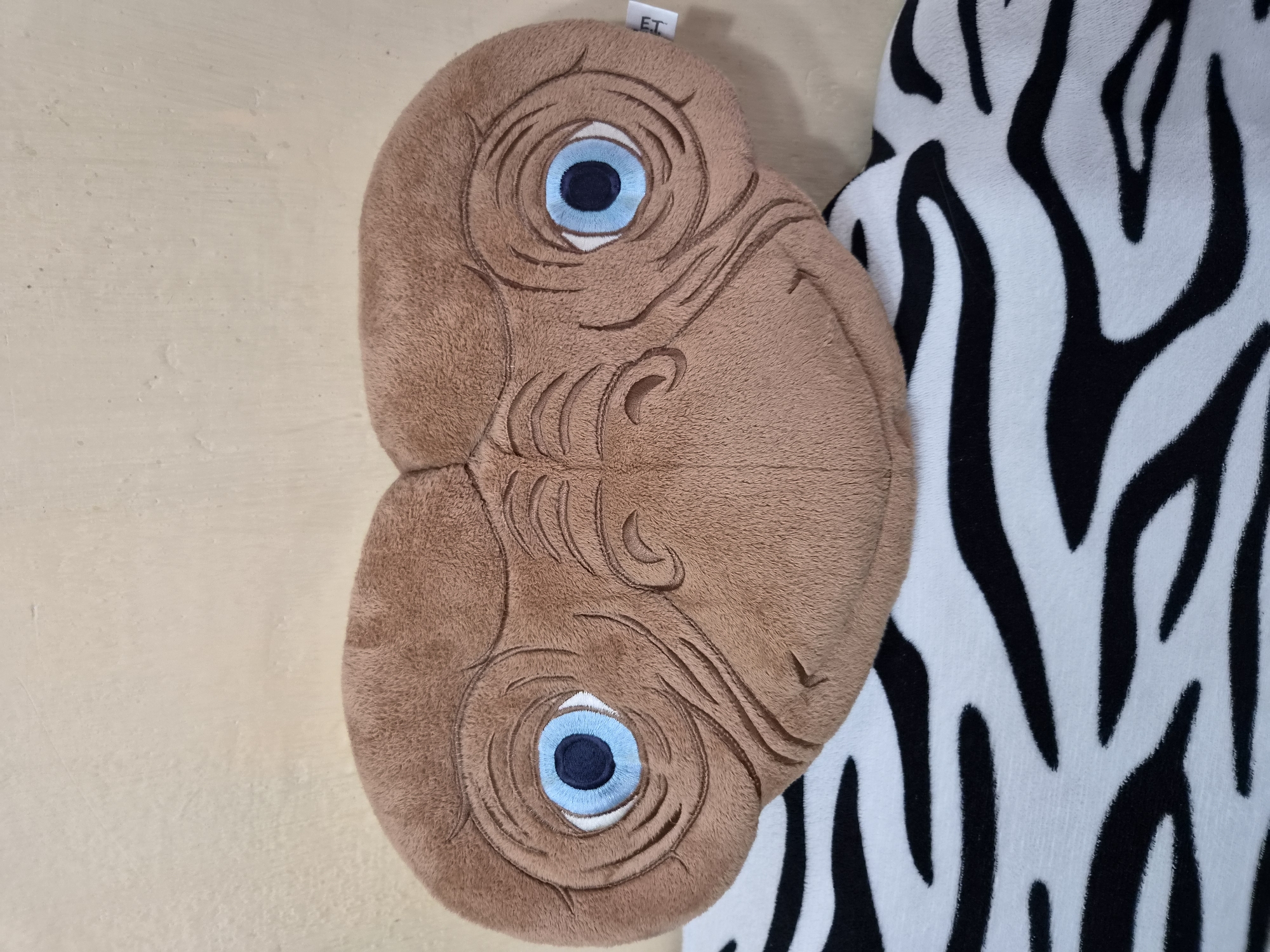 A plush toy resembling the character E.T. is positioned on top of a zebra-striped blanket. The toy, made of brown fuzzy material, features large, blue-rimmed eyes with dark pupils, and sculpted facial wrinkles. The background consists of a textured, light-colored wall. A small white tag with the letters "ET" is visible on the right side of the toy. The image was captured in Arseniev, Russia.