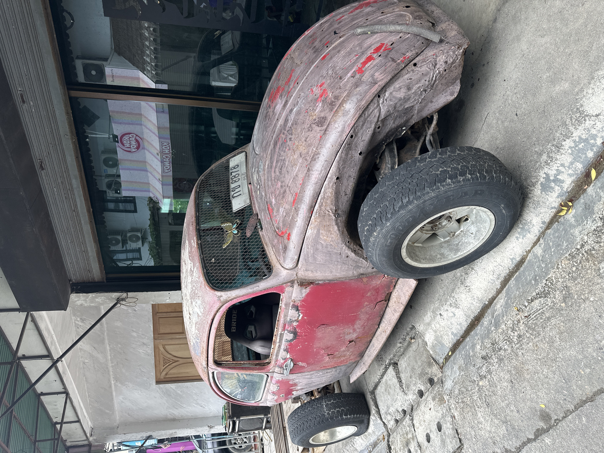 An extensively modified, heavily rusted red Volkswagen Beetle, styled as a rat rod, is parked on a concrete sidewalk in ตำบลเกาะพะงัน, Thailand. The vehicle exhibits significant paint loss and rust across its body panels, revealing exposed metal and patches of original red paint. The fenders are absent, exposing the suspension and large, white-rimmed wheels with robust tires. The interior features a black "BRIDE" racing seat visible through the open driver's side window. Both the windshield and the side windows are covered with a protective metal mesh, and a decorative blue butterfly is affixed to the front mesh. A Thai license plate, "กถ 8878", is mounted on the front hood.
The car is positioned in front of two commercial buildings. To the left, a building presents a light grey, textured stucco facade with a prominent pair of carved wooden double doors. Above this section, an industrial-style metal awning frame with a green tarp is visible. To the right, a storefront features large glass panels reflecting the interior, which includes two white air conditioning units, and a reversed reflection of a colorful awning displaying "KOH CANDY". The ground is composed of textured concrete pavers and a narrow drainage channel, with a few yellow leaves scattered near the front right wheel of the Beetle. In the background, to the left of the car, a black speaker or utility box and the wheel of another vehicle are partially visible. The reflections in the glass storefront also show outlines of other vehicles and interior elements of the shop.