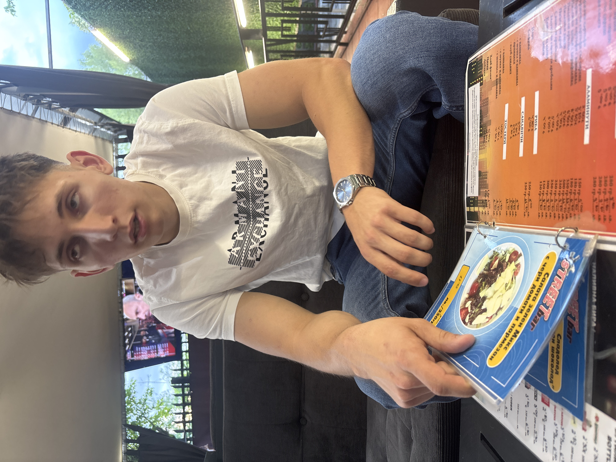 A young adult male, appearing in his late teens or early twenties with short, light brown hair, is seated on a dark grey tufted sofa, engrossed in reviewing a menu. He wears a white short-sleeved t-shirt with "ARMANI EXCHANGE" printed in black on the chest, blue denim jeans, and a silver-toned wristwatch on his left wrist. His gaze is directed upwards and slightly to his right, with his mouth slightly open. His left hand holds open a laminated menu displaying a blue-bordered page with an image of a salad and Cyrillic text, including "САЛАТА ЗЕЛЕН МИКС с чери домати и пармезан" (Green Mix Salad with cherry tomatoes and parmesan) and a price of "9 лв. / 250гр." A visible orange-toned page on the right, also in Cyrillic, lists categories such as "ТОСТЕРИ" (Toasts), "САНДВИЧИ" (Sandwiches), "РИБА" (Fish), and "АЛАМИНУТИ" (Quick Meals), indicating a cafe or restaurant setting.
Behind the male, to the left, a television screen displays a male figure in a suit, possibly from a broadcast, framed by dark curtains and visible greenery outside. To the male's right, beyond a dark railing, is an outdoor area with lush green foliage and bright overhead lighting fixtures, indicating a covered outdoor terrace or patio. The visible currency ("лв." - Bulgarian Lev) and Cyrillic script on the menu confirm the location as Bulgaria. This scene depicts a casual eatery, consistent with a street bar or cafe in an urban setting such as Blagoevgrad, Bulgaria.