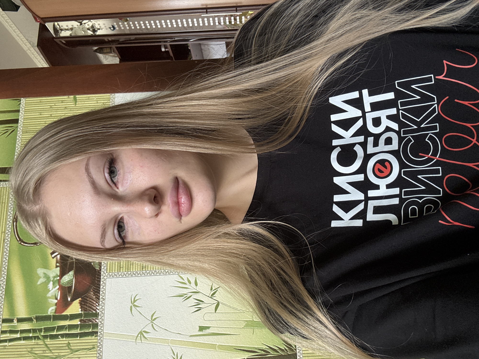 A young woman with blonde hair is pictured in a close-up shot, facing the camera. She is wearing a black t-shirt with white and red text printed on it. The background features a wall with a bamboo-themed wallpaper and a wooden cabinet on the right side. The woman's face is slightly tilted, and her hair is flowing around her shoulders. The lighting suggests an indoor setting. The location specified, Комарихинский, Russia, is noted, but there is no visual information in the image to confirm or deny this specific location. The image is a portrait of the woman.
