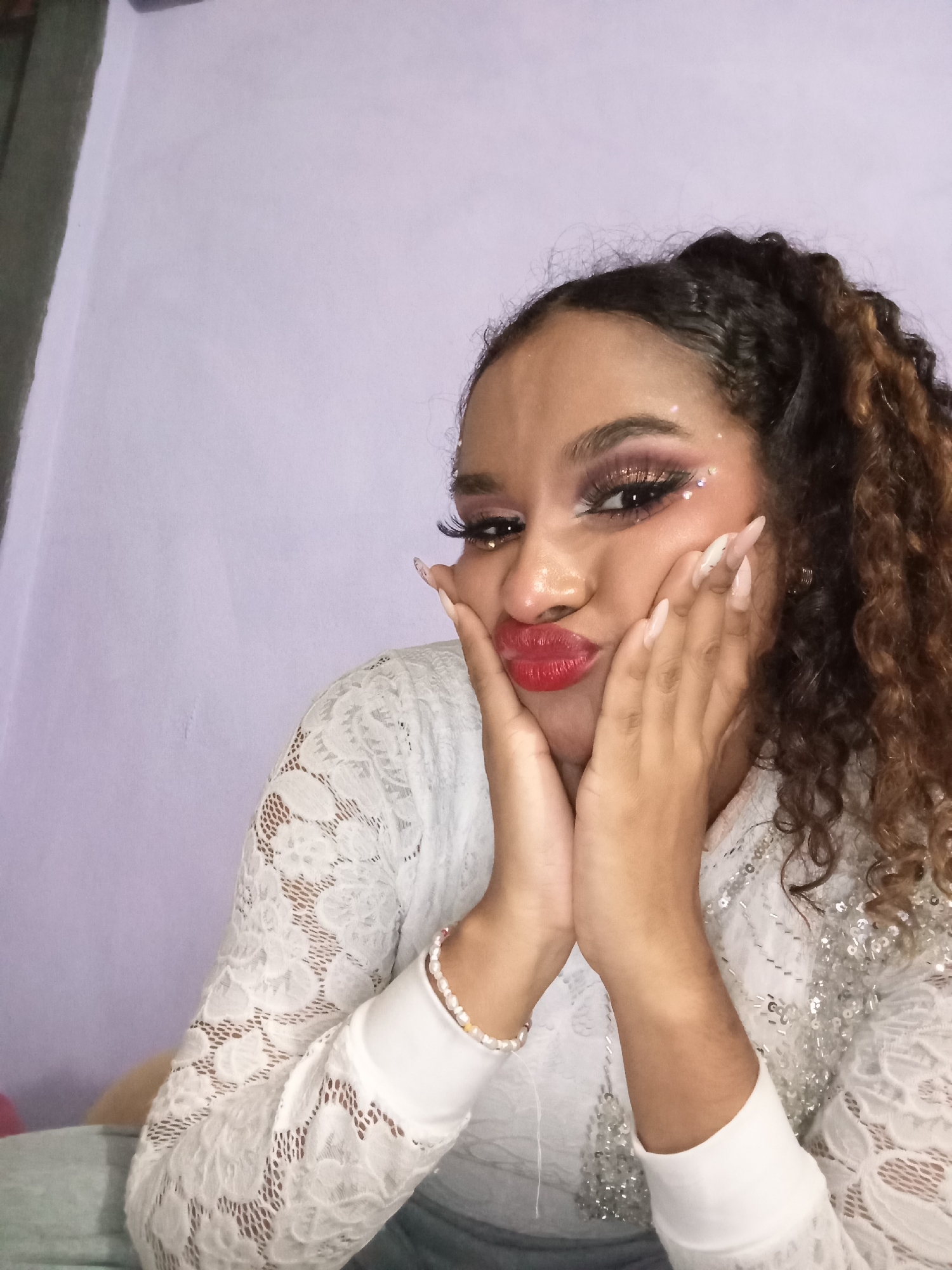A young woman is posing for a selfie. She is looking at the camera and puckering her lips into a kissy face while holding her cheeks with her hands. She has brown curly hair with caramel highlights pulled into a high ponytail. She is wearing elaborate makeup, including false eyelashes, glittery eyeshadow, and red lipstick, and her nails are long and painted light pink. She wears a white lace top with a white cuff and is wearing a pearl bracelet. The background is a light lavender wall. There are no indications of the location of Juan Griego, Venezuela.