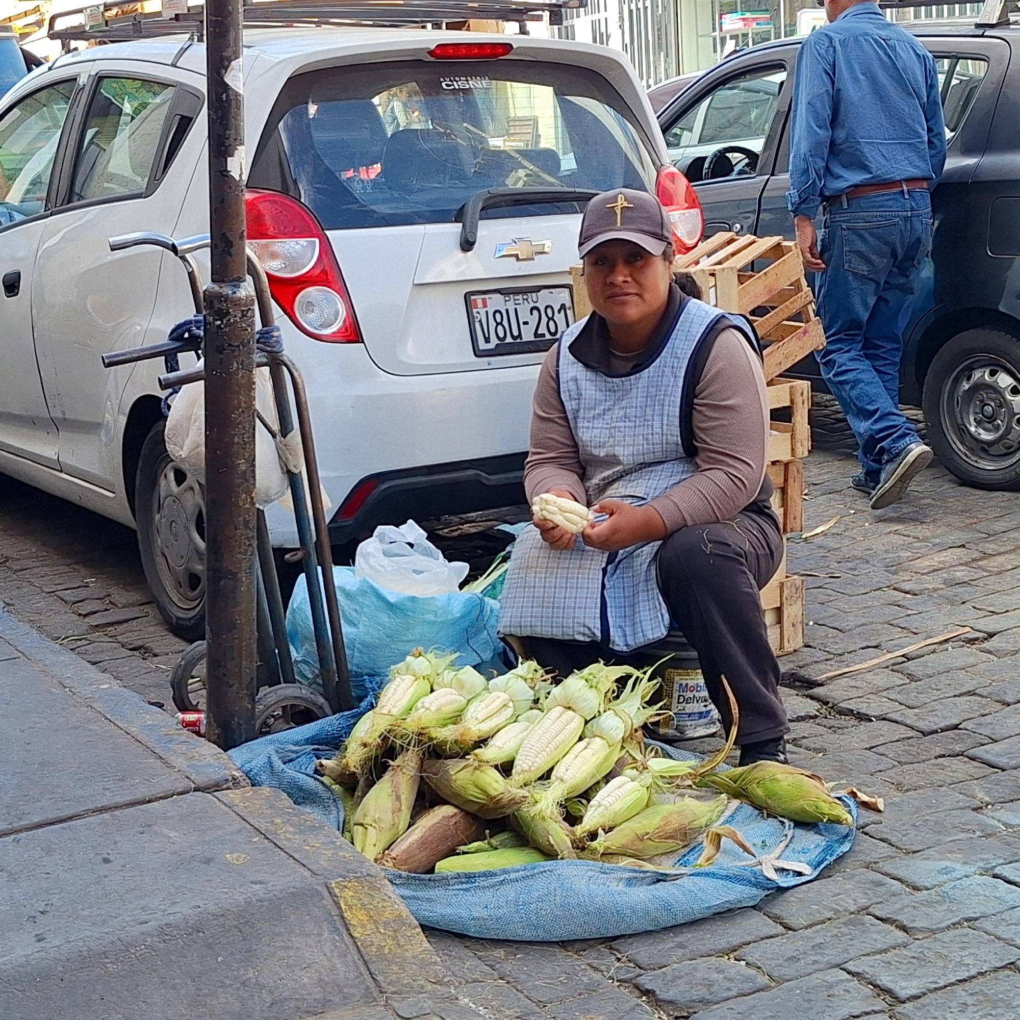 A woman is sitting on wooden crates on a brick sidewalk next to a pile of corn on a blue tarp in the Unknown, Unknown. The woman is wearing a brown long-sleeve shirt and a blue-and-white apron and is holding a peeled ear of corn in her hands. She is also wearing a dark-colored baseball cap with a golden cross. Behind her, a white car is parked with a Peruvian license plate, and a man in a blue shirt and jeans is walking toward another car. A metal pole and a small cart are situated near the corn and woman.