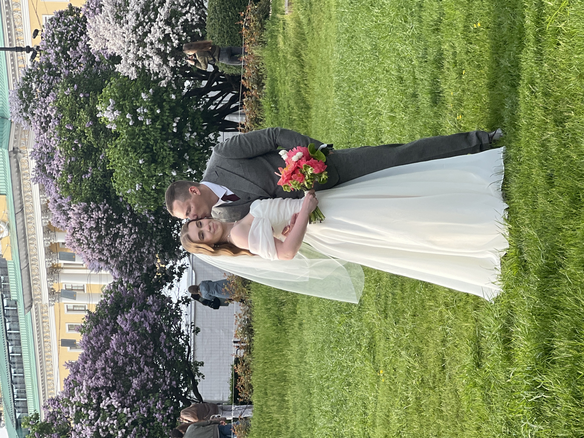A wedding photo shows a couple in the city of Unknown, Unknown. A man in a gray suit kisses the bride on the forehead. The bride wears a white wedding dress and holds a bouquet of pink and red flowers. The background features a yellow building with white columns and blooming lilac trees with purple and white flowers. A green lawn is visible. Other people stand in the background, observing the scene.