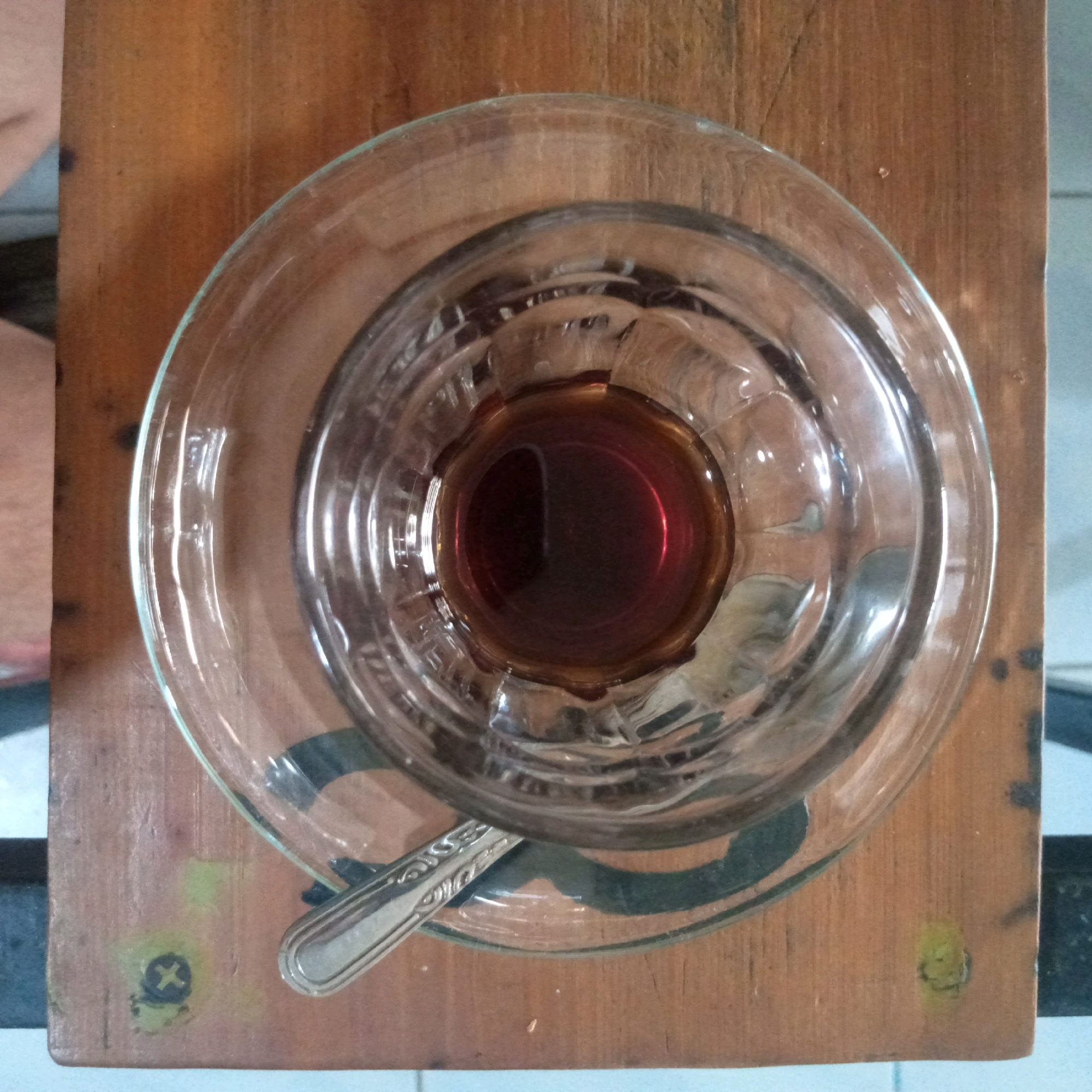 A top-down shot shows a clear glass cup filled with dark brown liquid, likely coffee, resting on a clear glass saucer. The saucer sits on a wooden surface, possibly a table, with visible wood grain. A silver spoon is placed diagonally across the saucer. The wooden surface has some minor wear and tear, including a yellow stain and a black circular marking with a white cross. The background is blurred, suggesting an indoor setting. The image captures a moment of stillness, with no people or overt actions depicted.