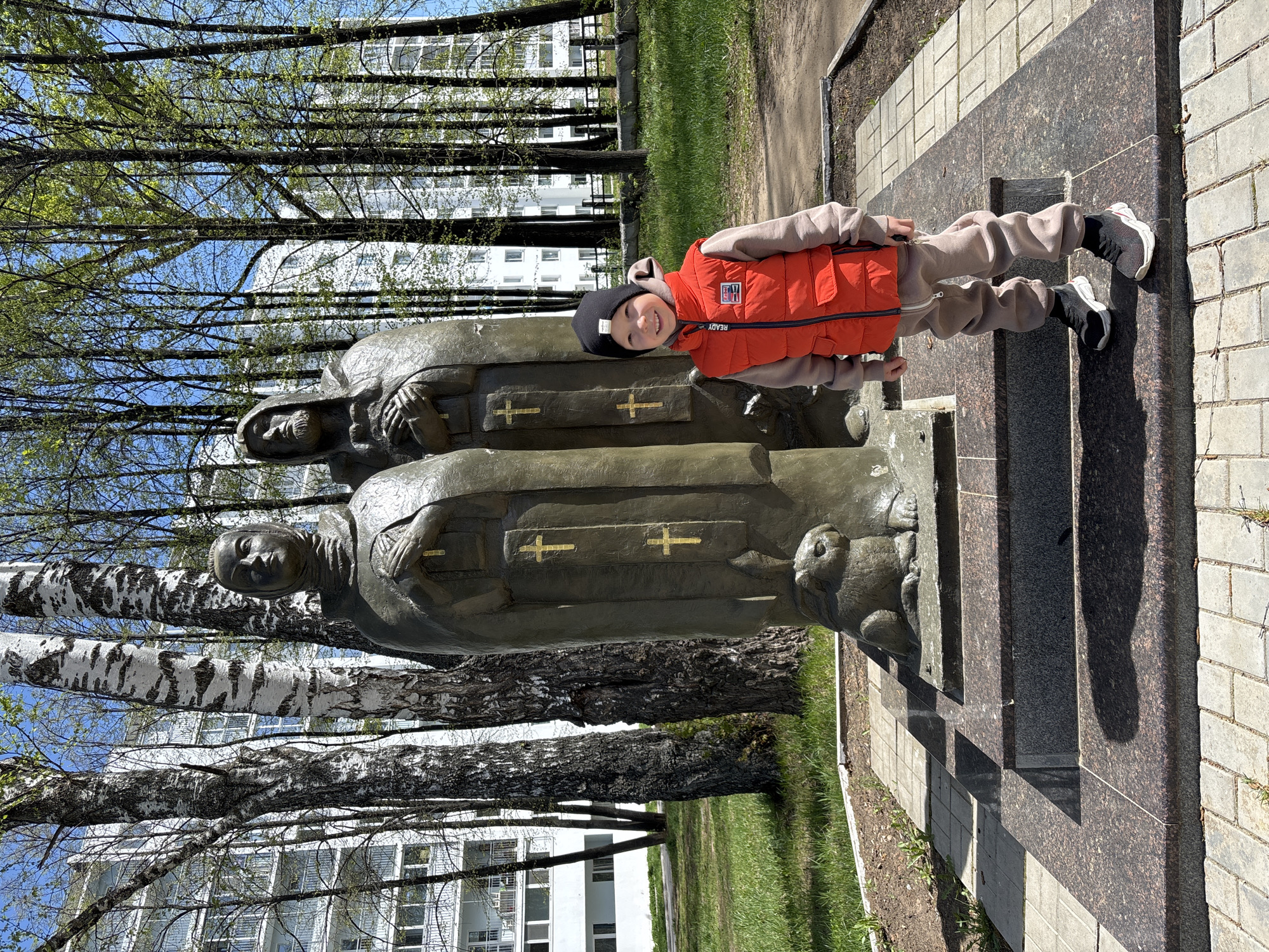 A full shot shows a young child standing in front of a statue of two women in Chaikovsky, Russia. The statue is a bronze color, and the women are wearing long robes with crosses on the front. The child is standing in front of the statue, facing the camera. He is wearing a black beanie, a beige sweatshirt and pants, and a red vest. The background includes birch trees and a white building. The statue stands on a concrete base covered in granite or similar stone. The statue and child are standing on a brick walkway and dirt area. The statue depicts nuns or religious women.