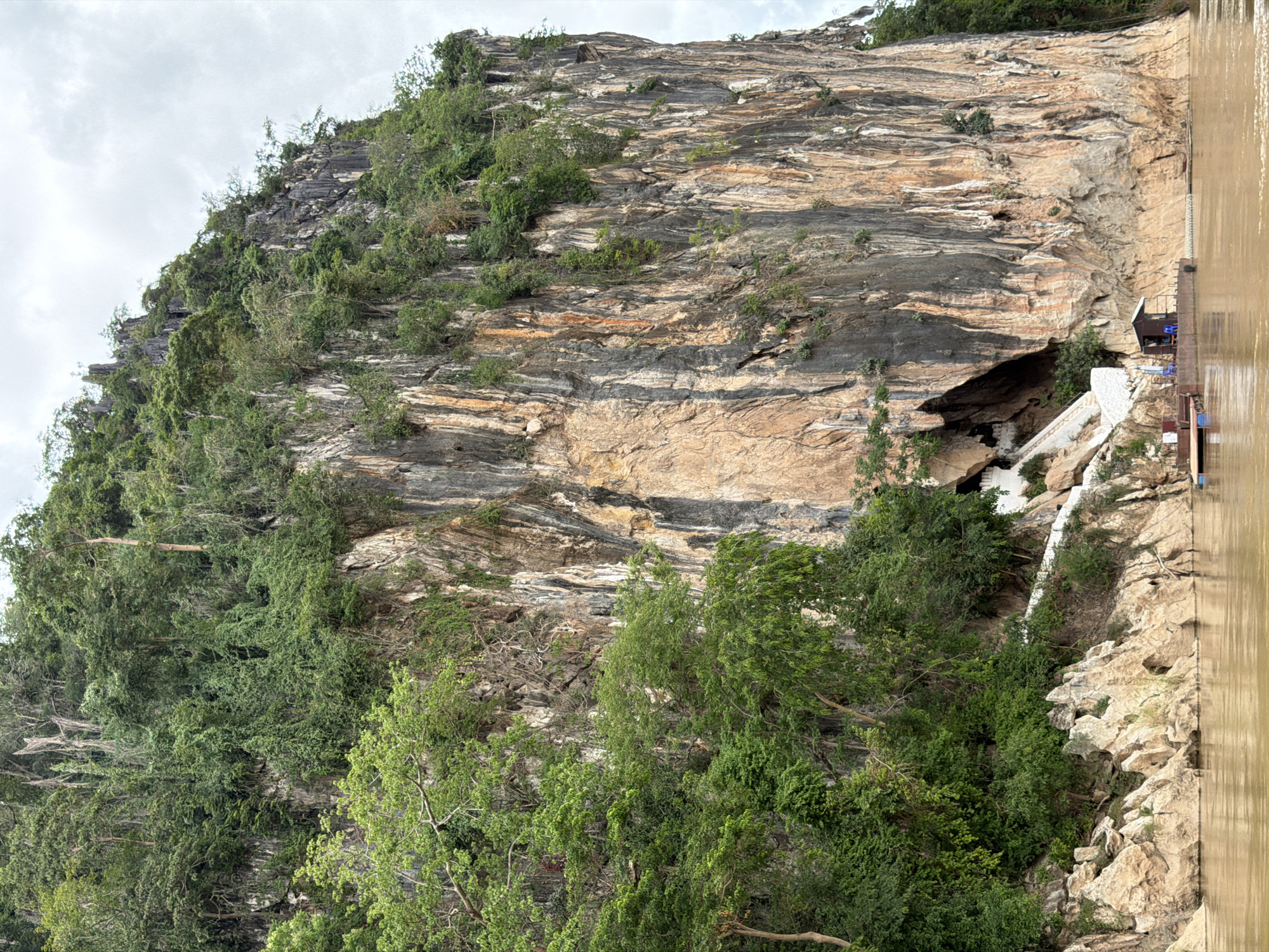 The image shows a massive, sheer cliff face covered in dense green vegetation near the city of Muangkeo, Laos. A muddy river flows in the foreground. At the base of the cliff, there is a small dock with a simple wooden structure, a few chairs, and a small boat. A white staircase leads up the cliff to a cave entrance, which is partially obscured by greenery. The sky is overcast.