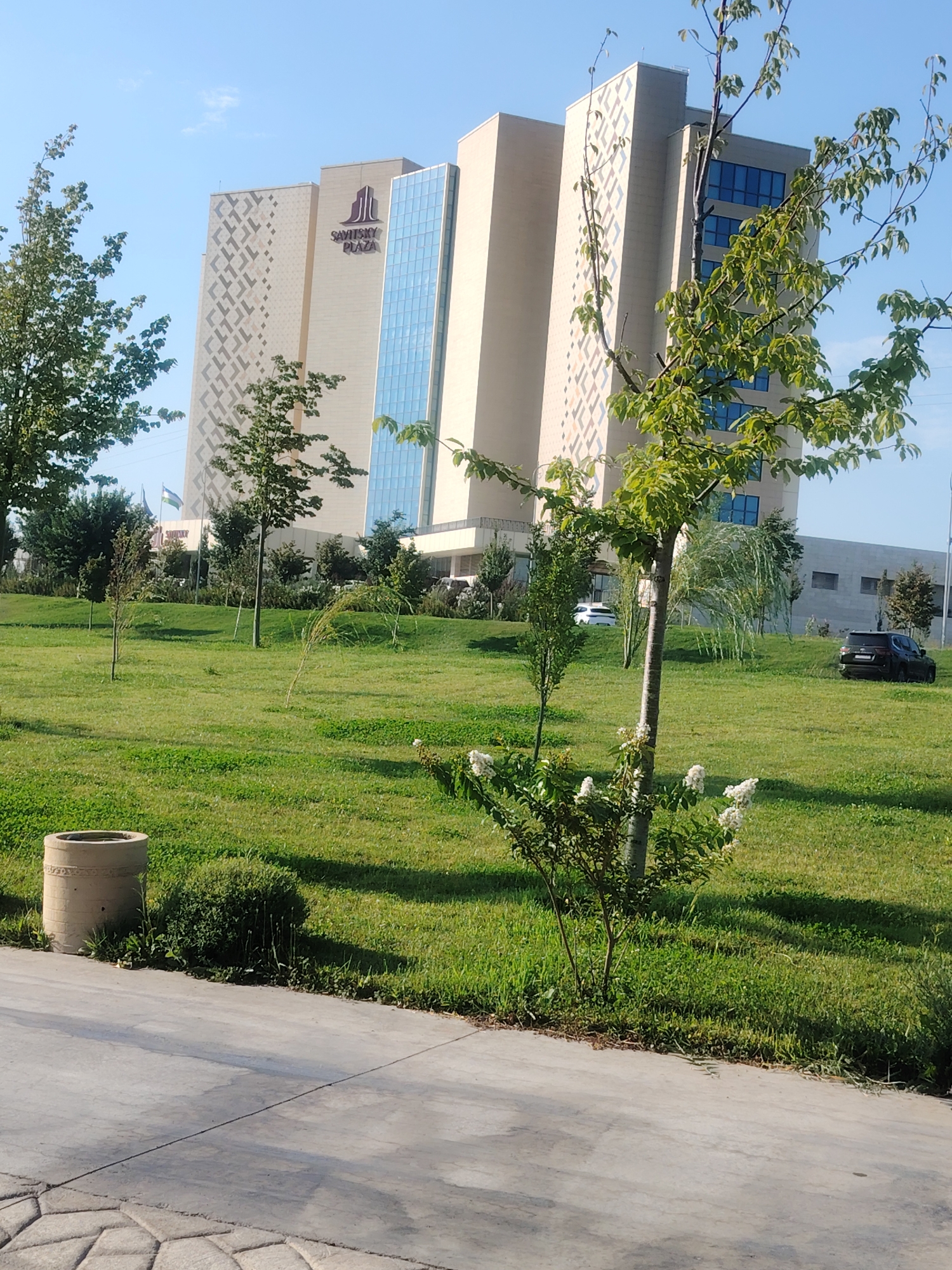 The image depicts the Sayidsky Plaza hotel in Konigil, Uzbekistan, on a clear, sunny day. A large, modern hotel building with a light-colored facade and geometric patterns stands in the background. The building features large blue-tinted windows. In the foreground, a lush green lawn is dotted with several trees and bushes. A small bush with white flowers is prominently featured in the middle ground. To the left, a concrete planter is visible. A light-colored car and a dark SUV are parked on the side of the road behind the lawn. The sky is a clear blue.