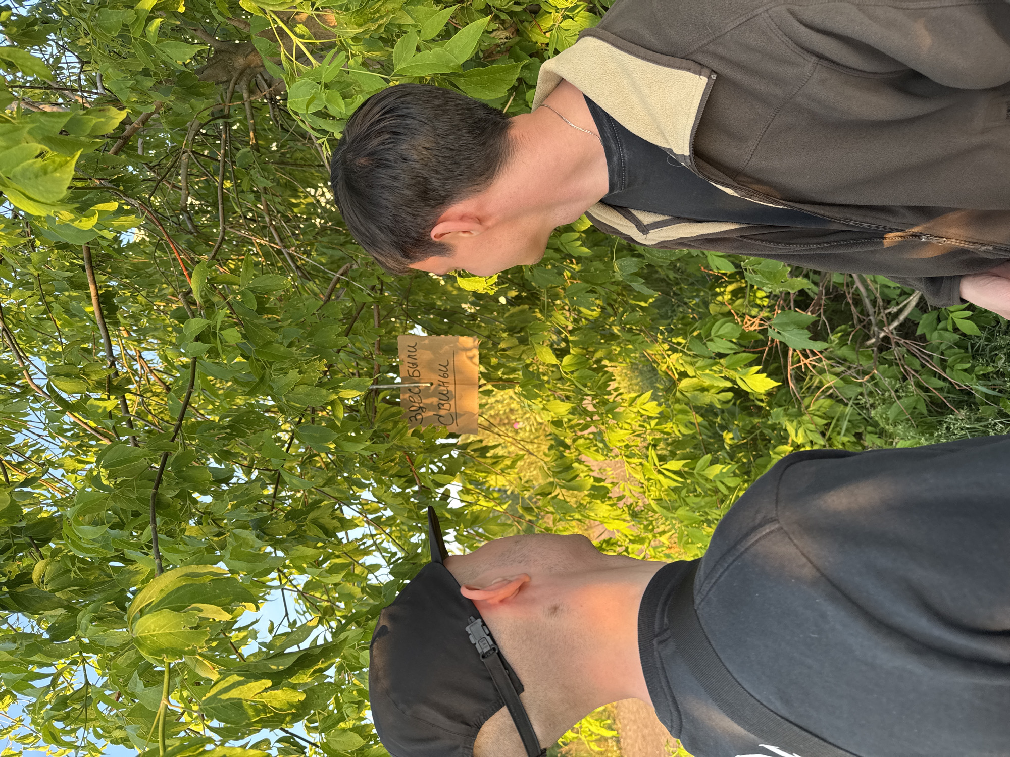In a natural setting with lush green foliage and a bright sky, two individuals are observed from behind, looking towards a handwritten sign on a tree. The sign, made of brown material, appears to have text written in a dark color. One person is wearing a black baseball cap and a black t-shirt, with a black strap visible over their shoulder. The other person is wearing a dark brown zip-up jacket with a light-colored collar over a dark t-shirt, and a thin chain necklace. The lighting suggests it is either early morning or late afternoon. The location is not identifiable as Saint Petersburg, Russia.