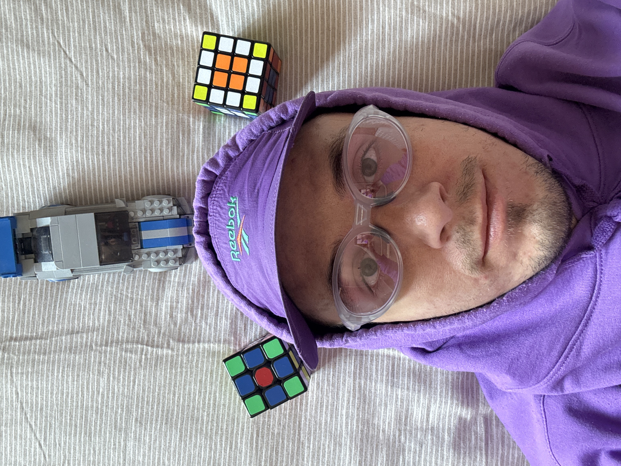 The image, taken from a top-down perspective, features a person wearing a purple hoodie and a purple cap with a "Reebok" logo. The person has on clear-rimmed glasses. To the left of the person's head is a Rubik's cube, and to the right is another, larger Rubik's cube. Above the person's head, positioned centrally, is a gray and blue Lego car. The scene appears to be indoors, on a surface with horizontal striped texture, consistent with a bed or a similar textile. There is no identifiable information within the image to suggest a location in Đà Nẵng, Vietnam.