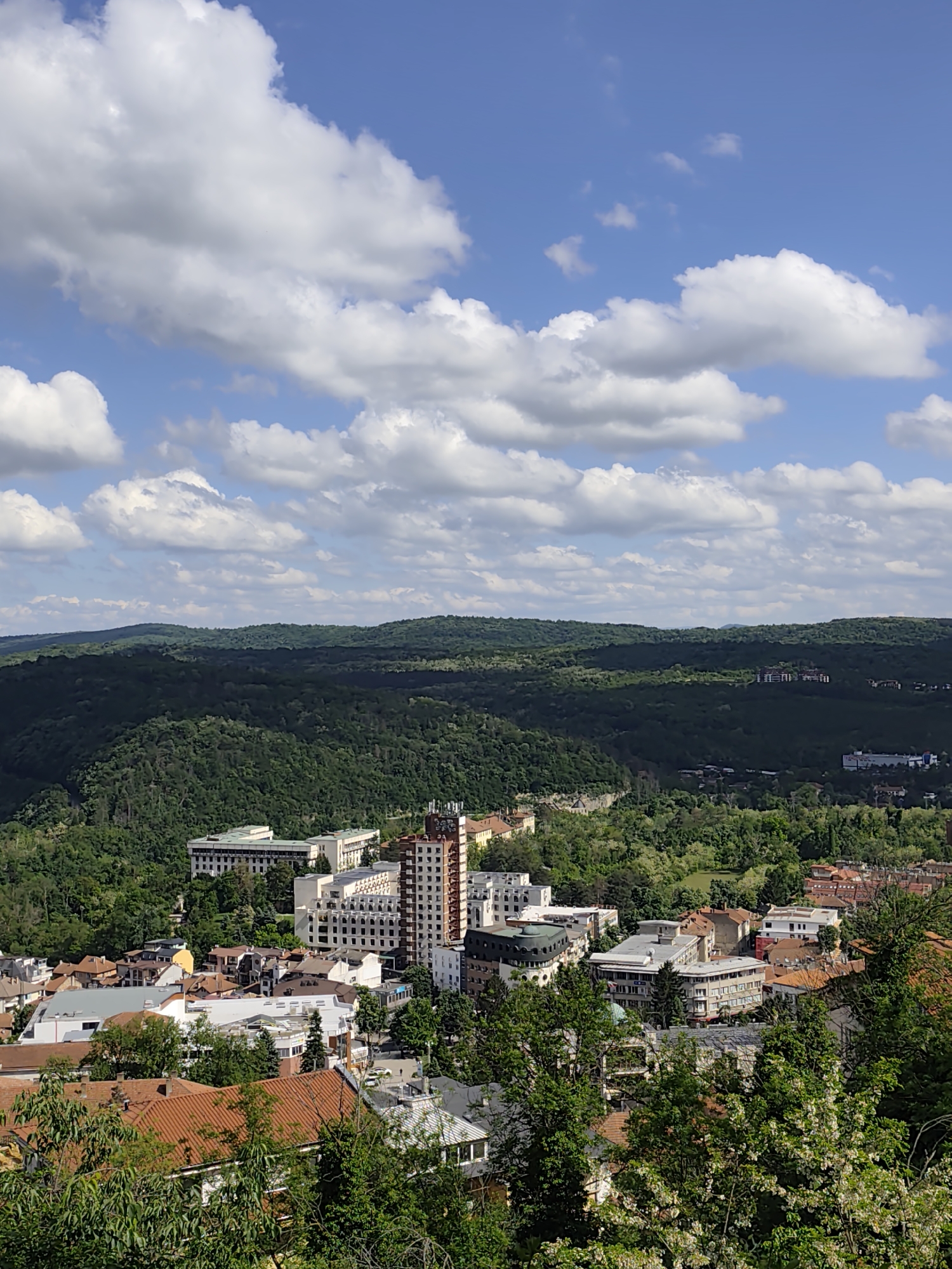The image shows a panoramic view of a city situated in a valley, surrounded by lush green hills and forests under a bright blue sky with scattered white clouds. In the foreground are buildings with red tile roofs mixed with trees. The architecture varies, including a tall building with a brick-patterned facade and white structures with a modern design. Behind the city, forested hills rise. The sky is clear and bright.