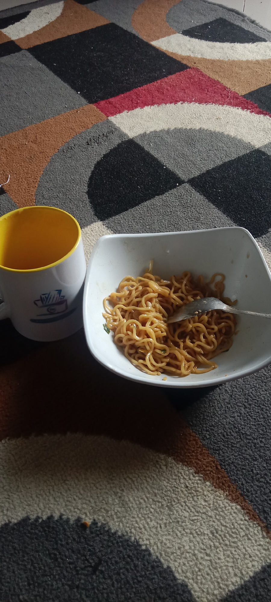 In a domestic setting in Krinjing, Indonesia, a white mug with a yellow interior and a blue logo sits to the left of a white bowl of noodles. A fork rests in the bowl, partially submerged in the noodles. The objects are placed on a carpet with a geometric pattern of grey, black, brown, and red. The lighting suggests it is daytime.