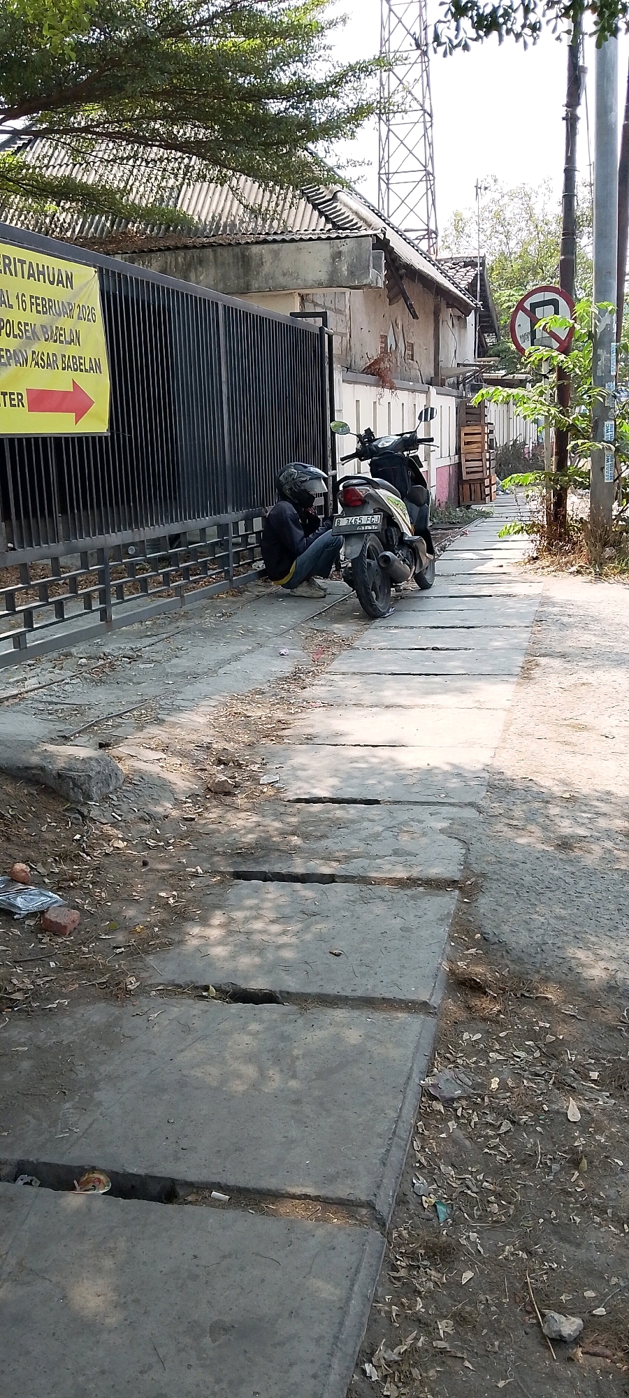 The image depicts an outdoor scene on a sunny day in Babelan, Indonesia. In the foreground, a person wearing a dark jacket and jeans, with a helmet on, is squatting next to a motorcycle. The motorcycle, a light-colored scooter, is parked on a paved walkway that appears to be made of concrete slabs. The person is leaning forward, possibly attending to the motorcycle. The walkway is bordered by a black metal fence on the left, behind which is a building with a corrugated metal roof. A yellow banner with Indonesian text and a red arrow is attached to the fence. Further back, a large cell tower is visible against a bright sky. To the right of the walkway, there is a dirt path with trees and foliage. A circular traffic sign with a red border and a diagonal red line across a bicycle symbol is visible on a pole. Debris, including fallen leaves, dirt, and some litter, is scattered on the ground. The overall impression is a gritty, urban environment with a focus on a man interacting with his vehicle in a public space.