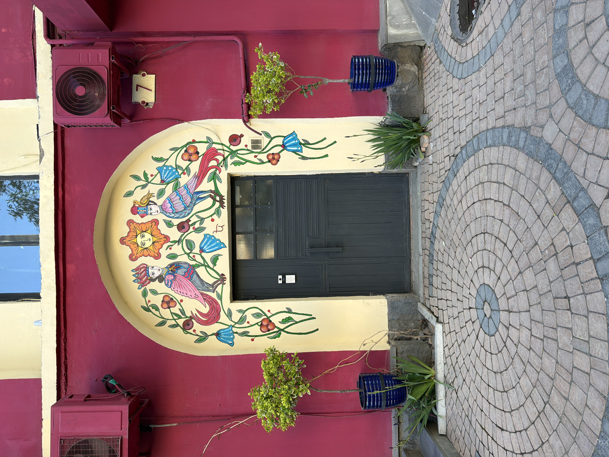 The image captures a vibrant building exterior, likely in Yerevan, Armenia, characterized by a bold color palette and decorative elements. The main wall is painted in a deep reddish-purple (magenta-like) color, contrasted with a cream-colored arched section above an entrance door.
A striking mural adorns the cream arch, featuring whimsical, stylized figures. It includes a central sun-like face, two anthropomorphic bird-like creatures with human heads (one crowned and another with flowing hair), along with blue bell-shaped flowers and green vines bearing red berries or fruits. The dark gray, vertically paneled double door below the mural has small windowpanes at the top and a small intercom/doorbell unit to its right.
Two reddish-purple outdoor air conditioning units are mounted on the left side of the wall, one near the top and another near the bottom. Above the upper AC unit, a small yellow plaque clearly displays the house number "7." Two decorative blue fluted pots, each containing a plant, are mounted on the wall to the right of the door, one above and one below the mural. Additional greenery is visible at ground level near the door.
The ground in front of the building is paved with light and dark gray cobblestones laid in an intricate pattern of concentric circles. A window on the far left reflects the bright sky, indicating daytime and clear weather conditions. The overall scene presents a static, colorful, and artistically decorated building facade.